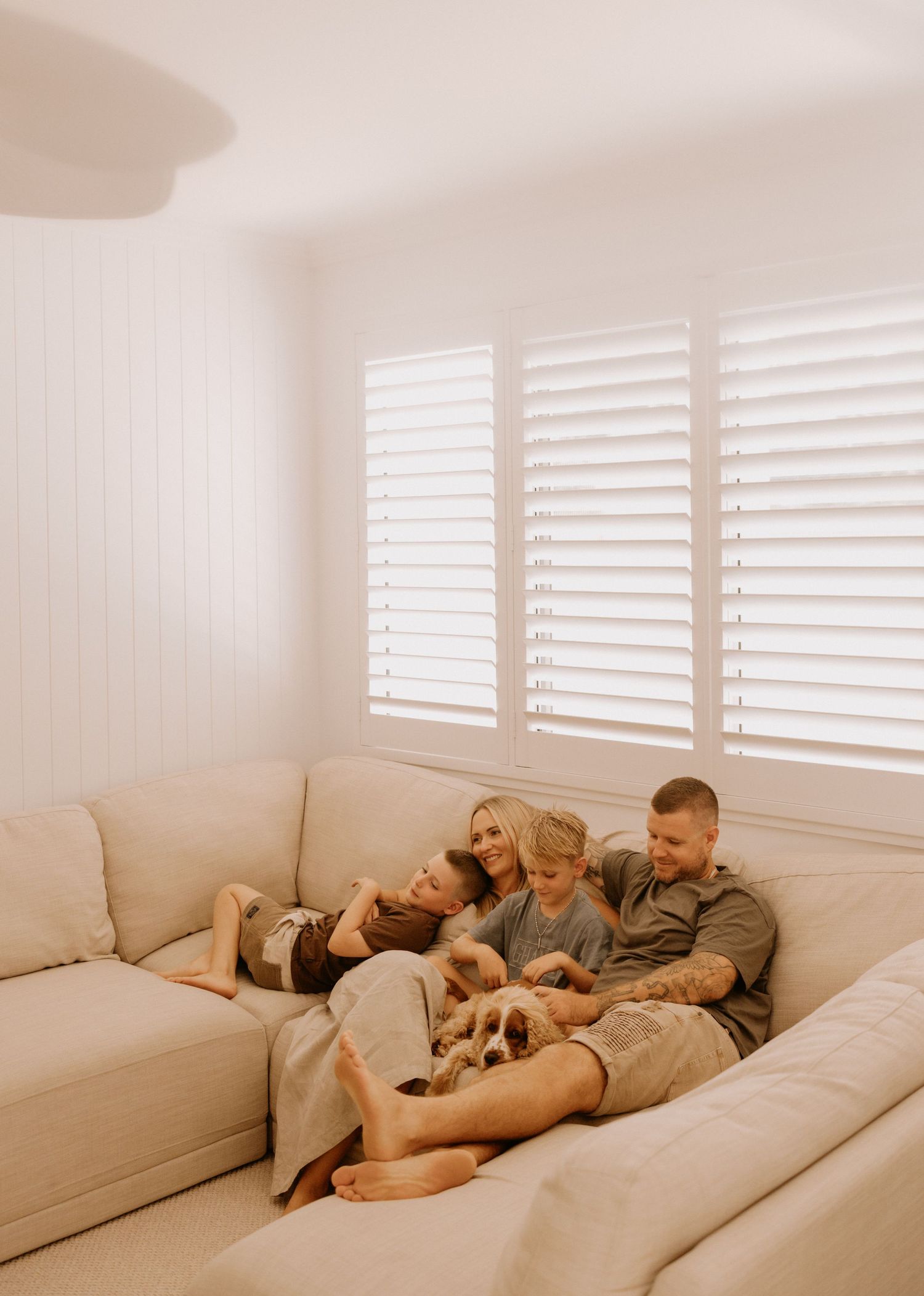 Family relaxes together on a cozy beige sectional sofa near white shuttered windows in a bright living room.