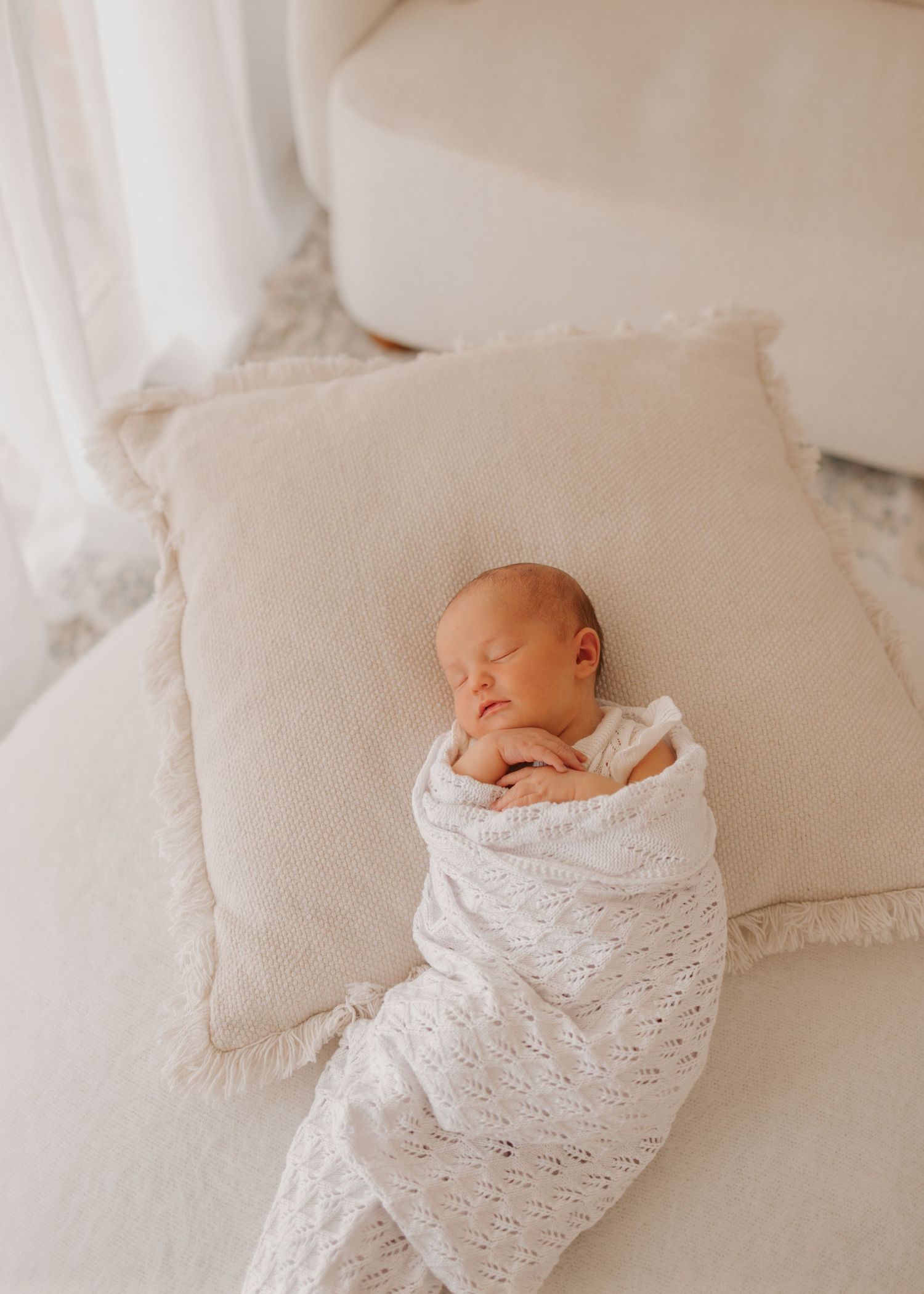 Peaceful sleeping newborn swaddled in white knit blanket resting on beige pillow against soft neutral background.