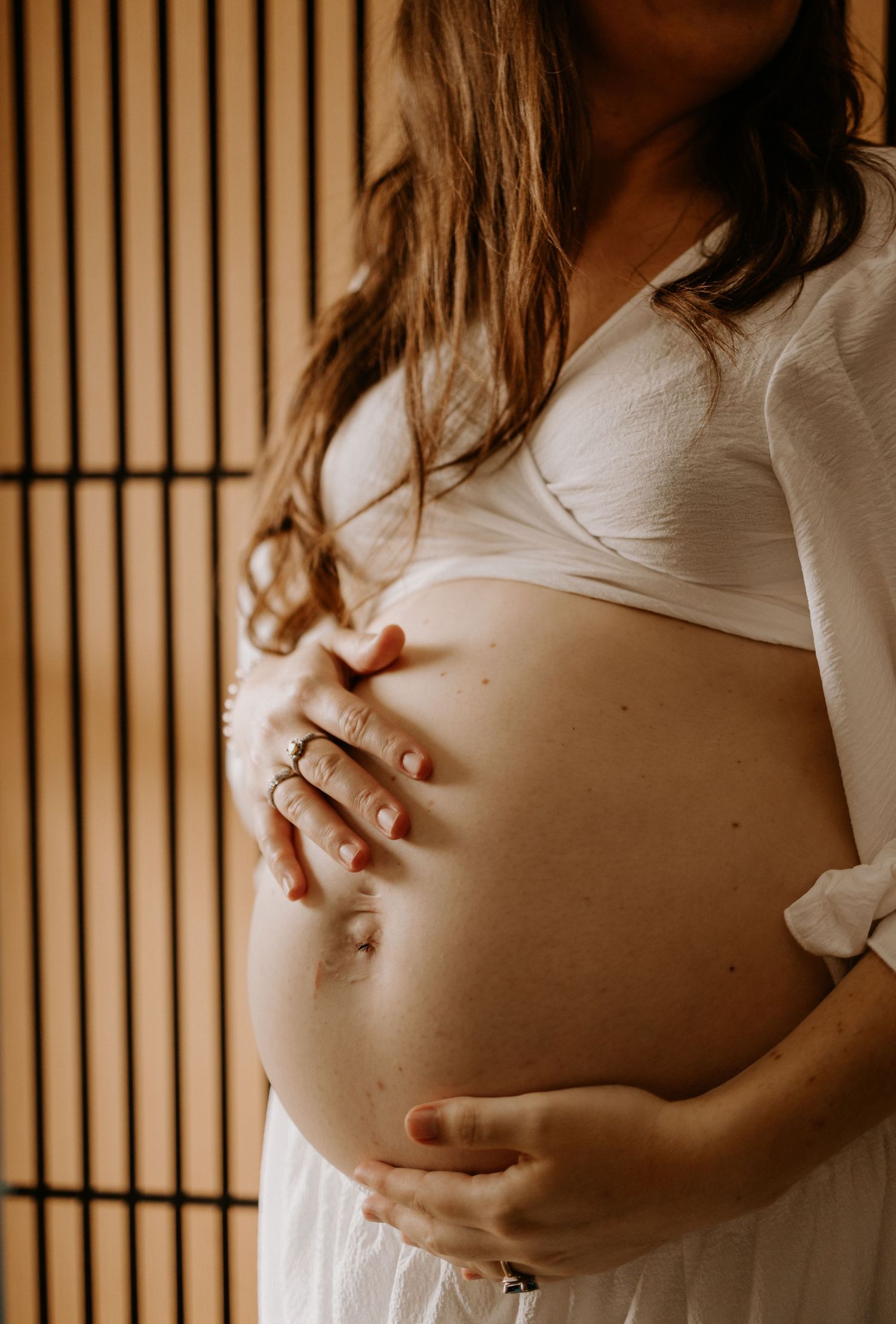 A pregnant woman in white clothing gently holds her baby bump against a wooden slatted background in warm lighting.