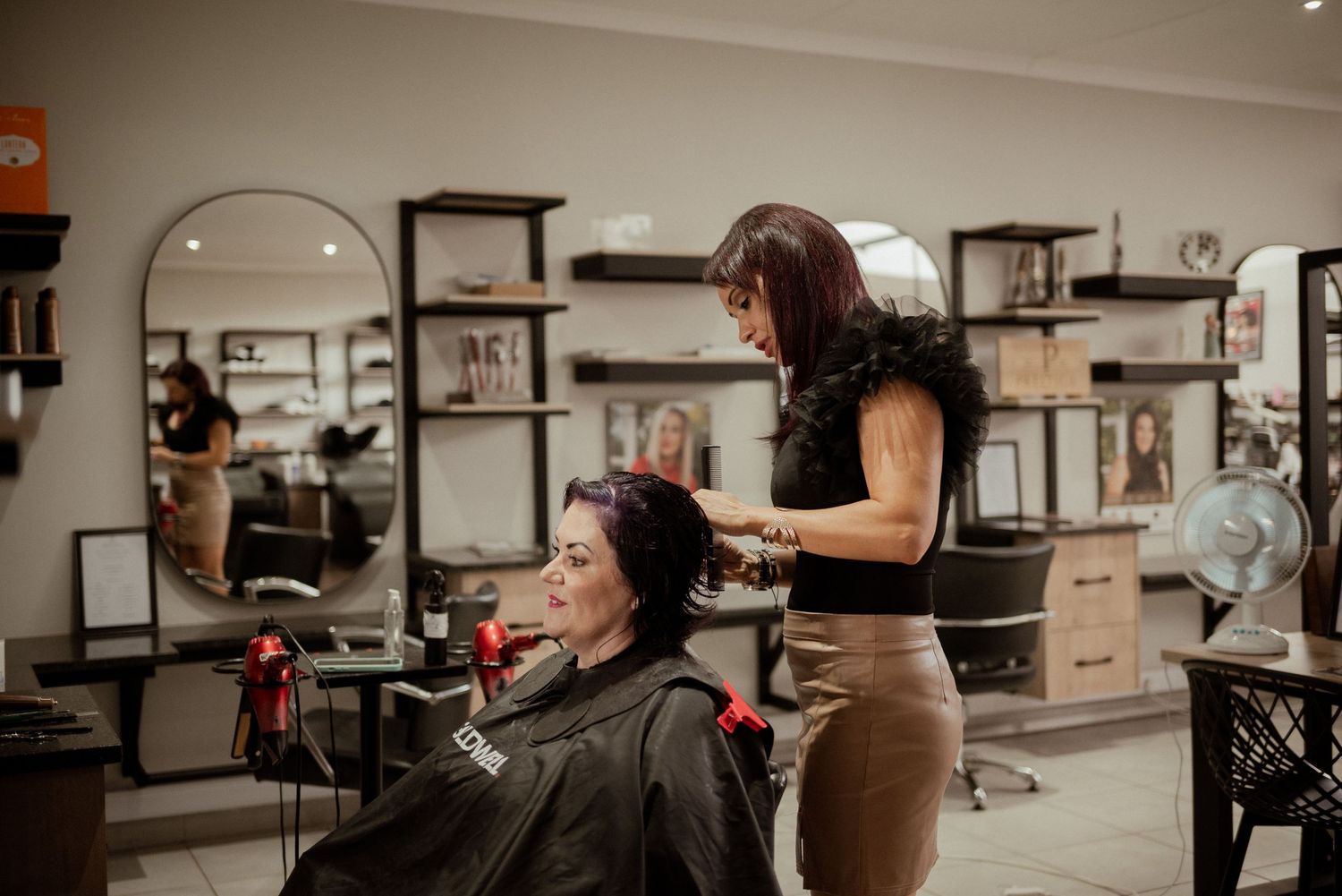 Professional hair stylist works on client's hair in modern salon with shelving displays.
