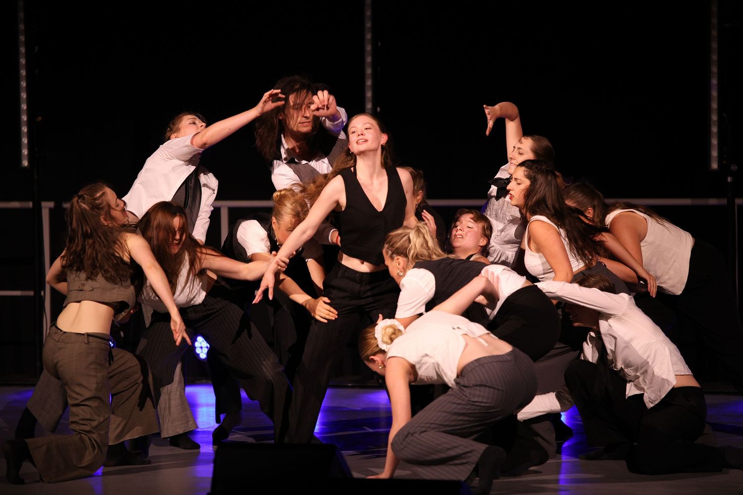 Performers in black and white costumes strike dramatic poses during an energetic dance number on a dark stage.