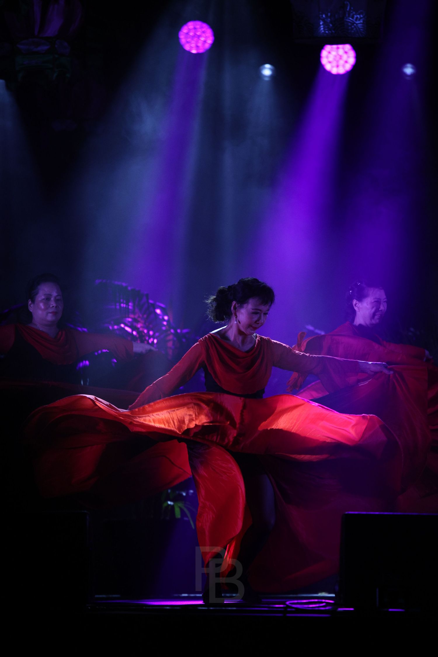 Dancers in flowing red costumes perform under dramatic purple stage lighting creating an ethereal atmosphere.