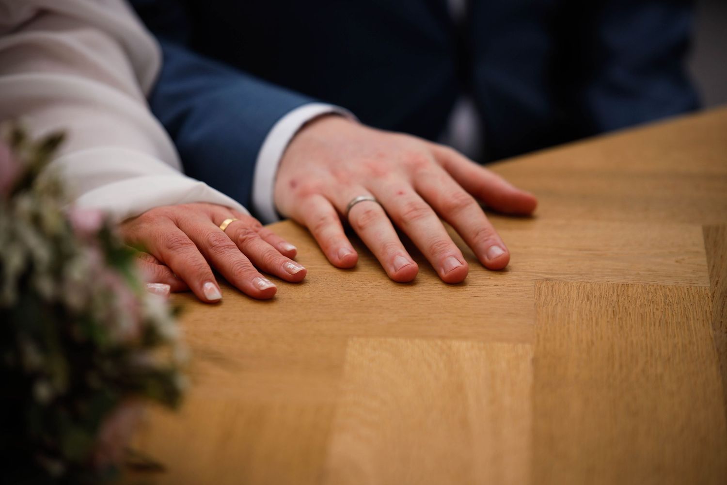 Two hands with wedding rings rest on a wooden table surface next to some greenery in the corner.