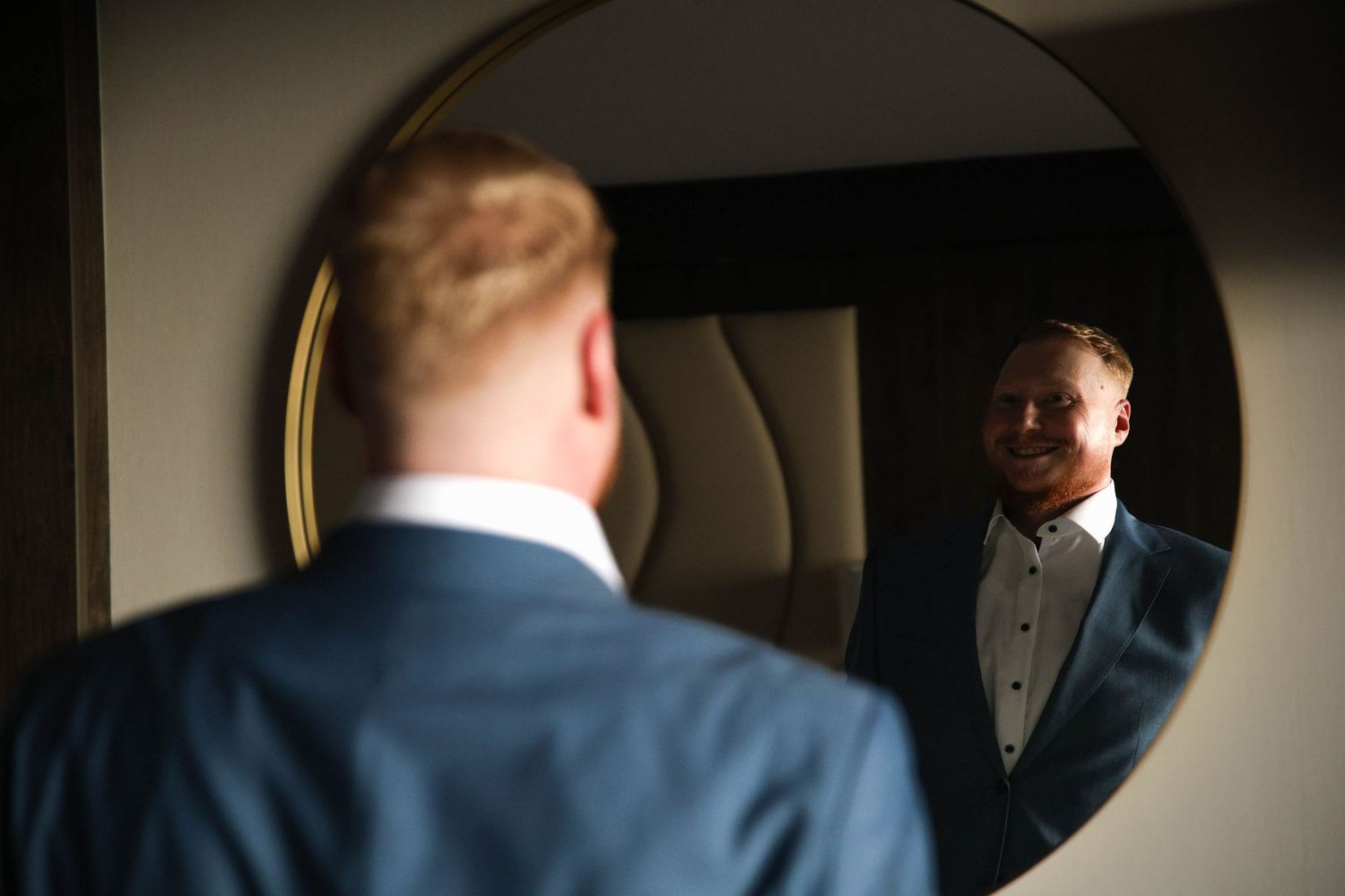 A person in a navy suit adjusts their tie while looking in a mirror.