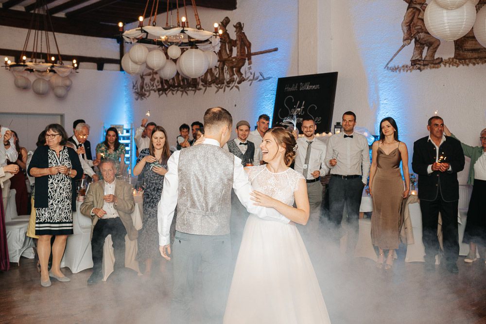 A couple shares their first dance while guests watch at a wedding reception with white foggy effects and rustic lighting.