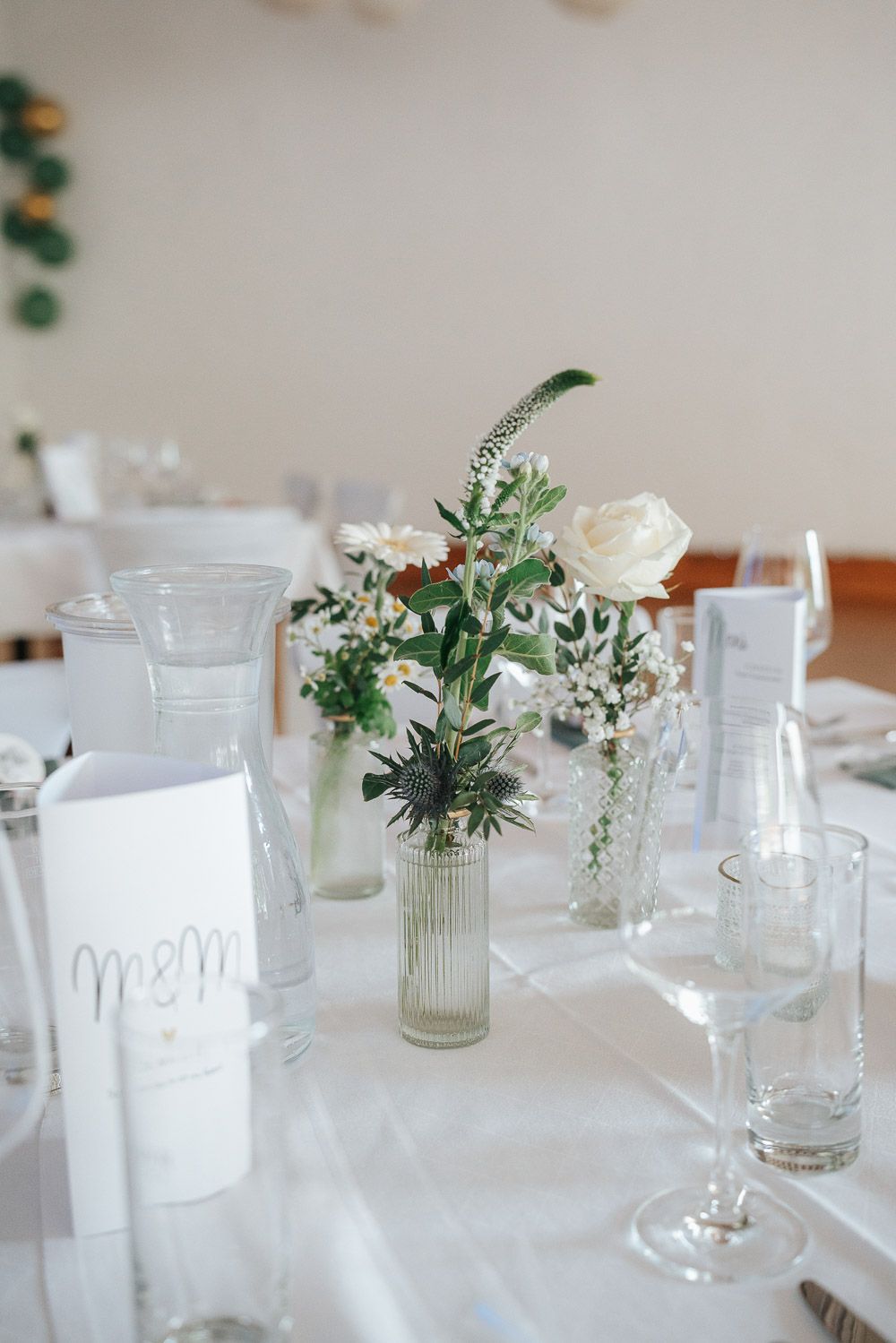 White table setting with delicate floral arrangements in glass vases featuring greenery and soft white blooms.