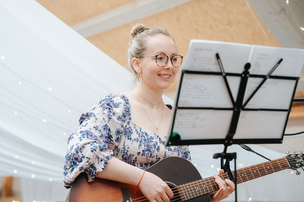 Musician in glasses wearing floral print dress performs with acoustic guitar at indoor venue with music stand.