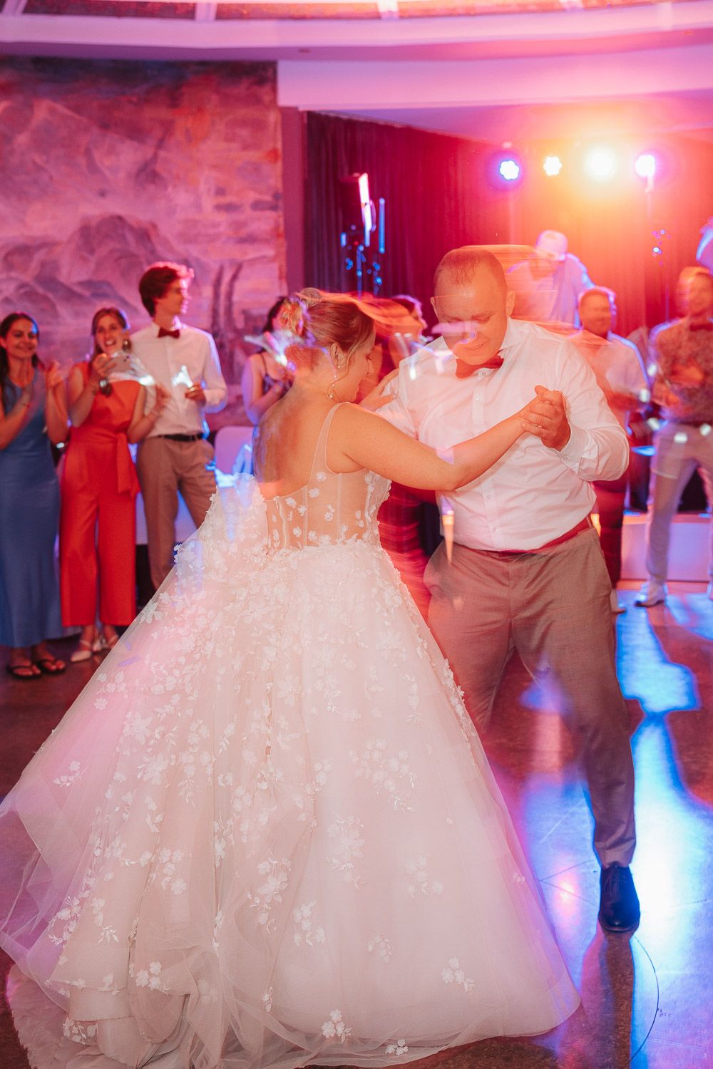 A traditional wedding dance scene with a bride in a sparkling white ball gown under romantic pink and red lighting.