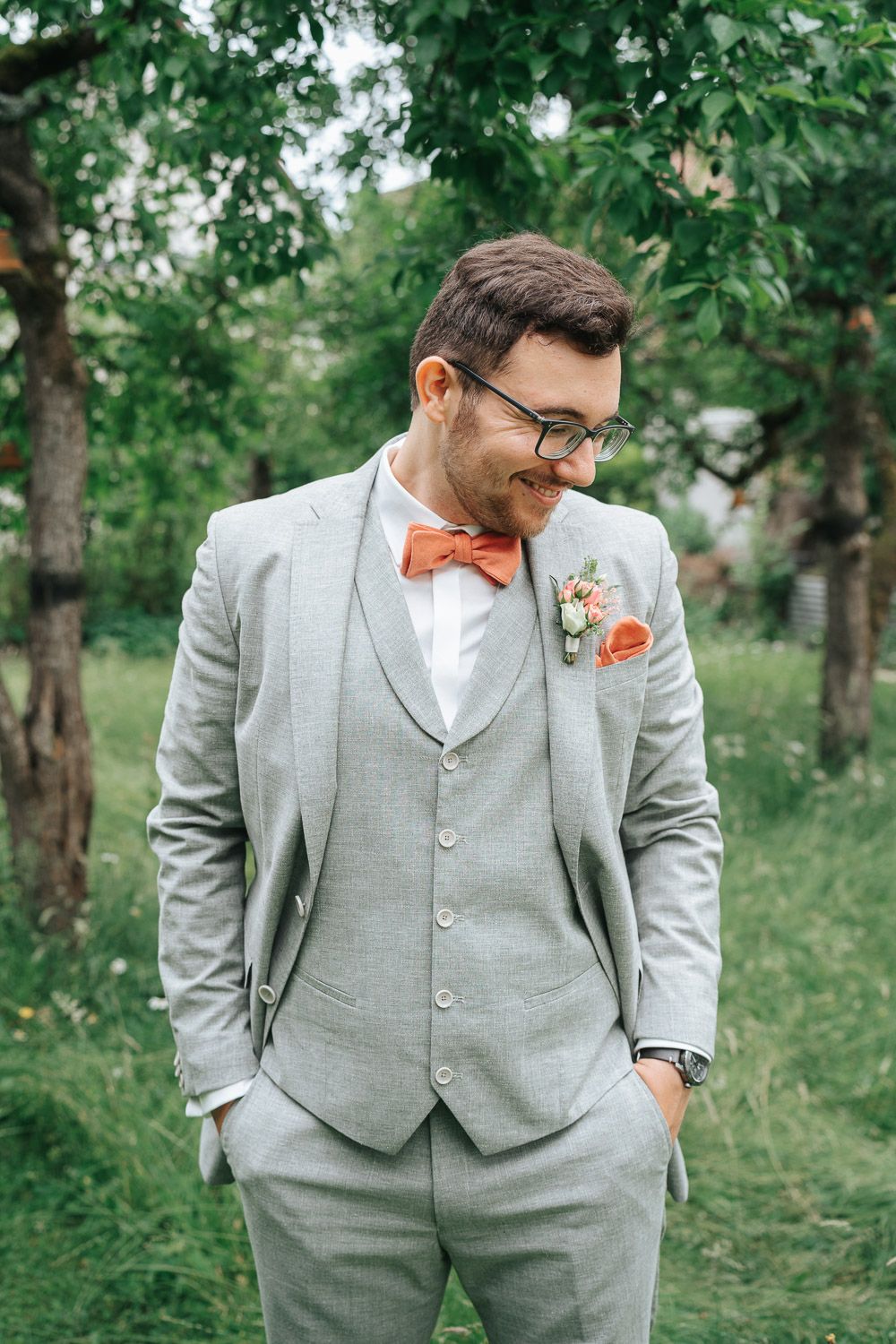 A groom wearing a light gray three-piece suit and orange bow tie poses outdoors in a garden setting.