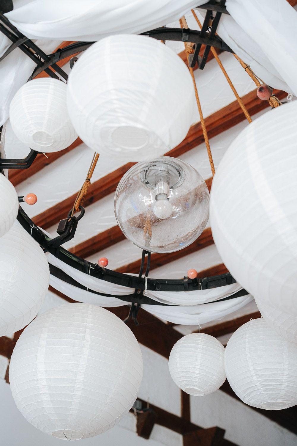 White paper lanterns and light fixtures hanging from wooden beams in an event space ceiling.