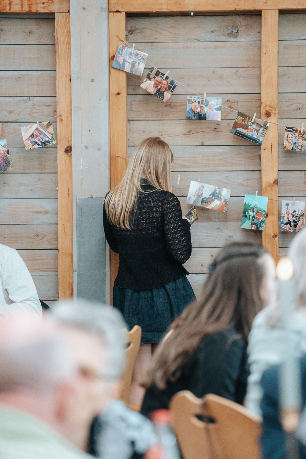 A person in black attire looks at photos displayed on wooden wall panels while others sit in chairs at an event.