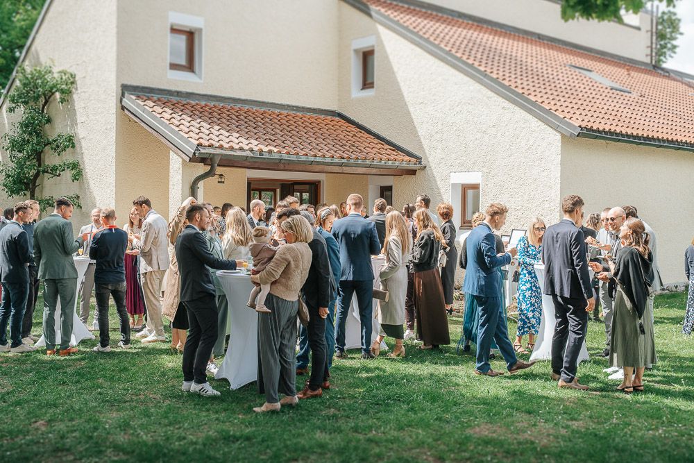 A group of formally dressed guests gather on a lawn outside a building with terra cotta roof tiles during a celebration.