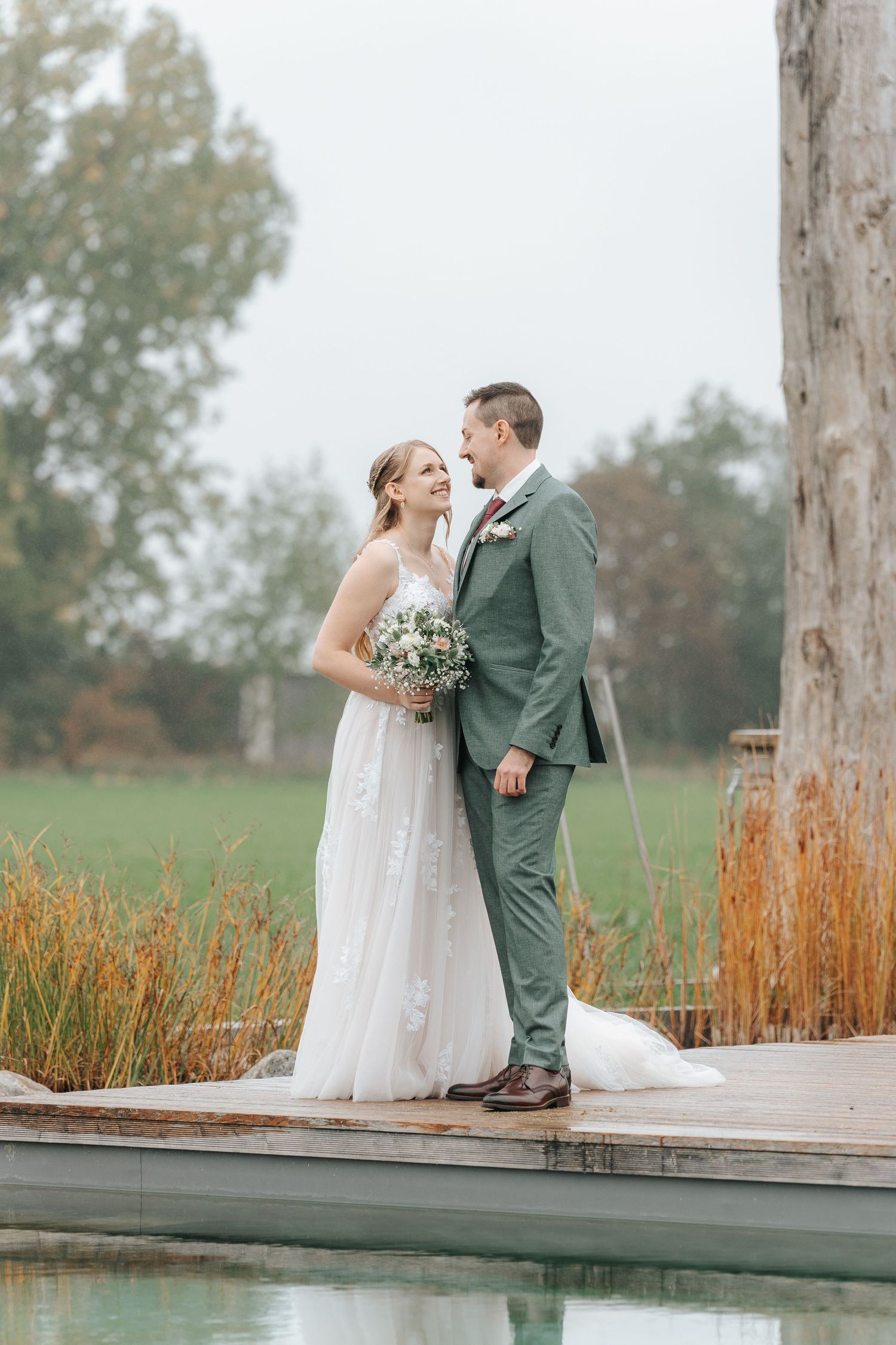 Newlywed couple shares romantic moments on a wooden dock surrounded by autumn grasses and misty scenery.