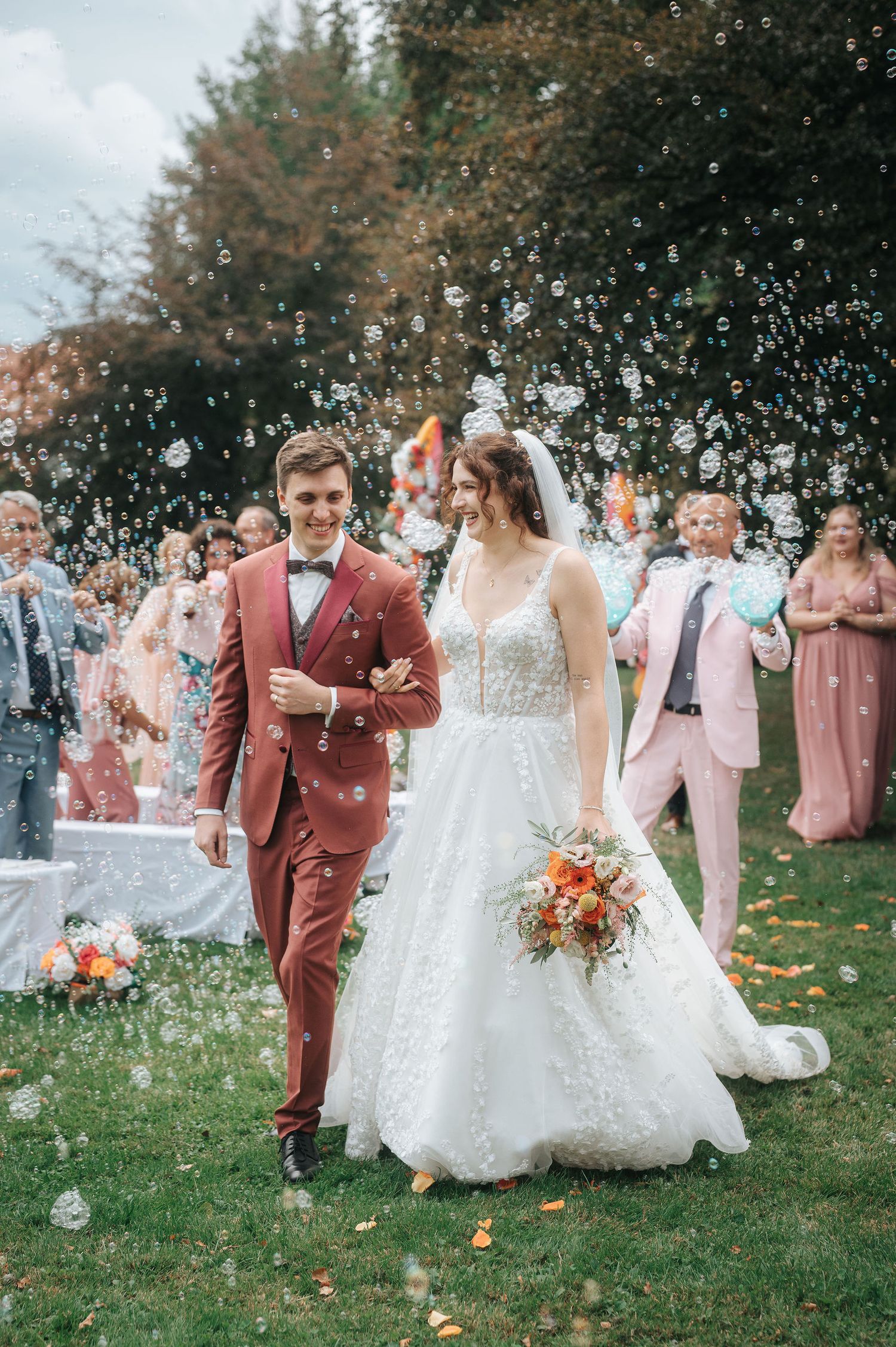 Bride in a lace wedding gown and groom in a rust suit walk arm in arm past cheering guests as bubbles fill the air at an outdoor wedding.