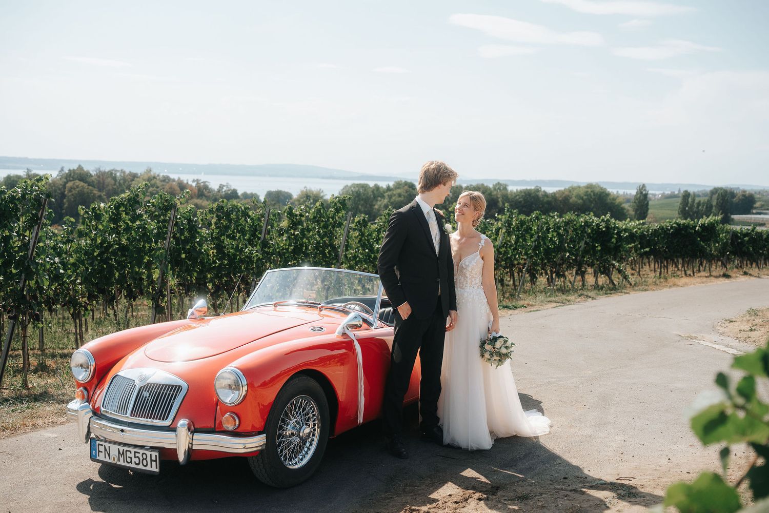 Bride and groom embrace beside a red vintage convertible on a gravel road with vineyards and a distant lake behind them.