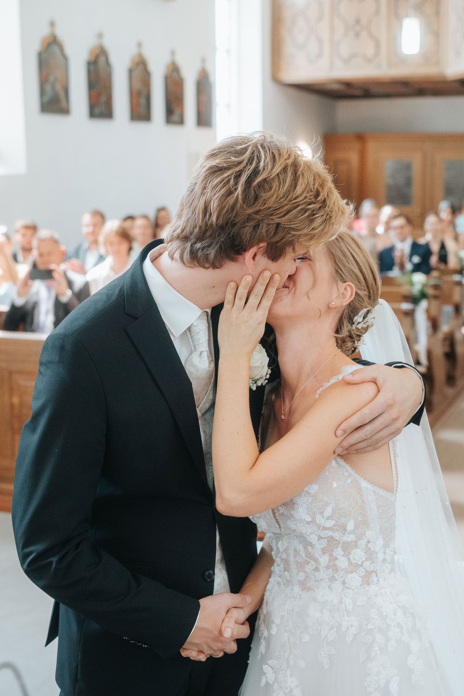 Bride in a lace wedding dress and veil shares a kiss with the groom in a dark suit during a church ceremony as guests watch.