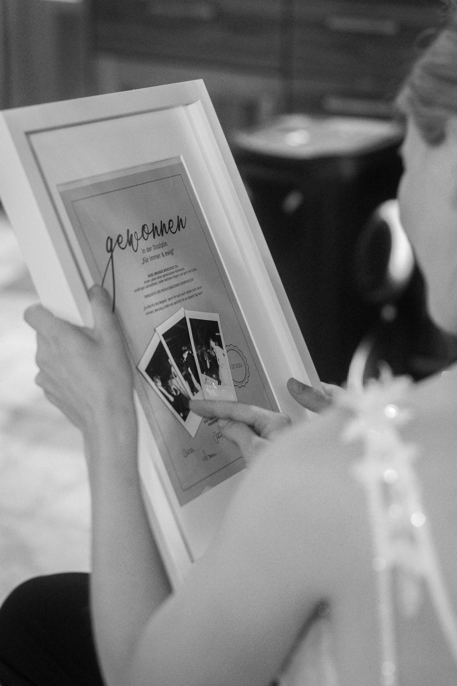 Black and white photo of a woman in a lace-trimmed dress holding a framed print with three small instant photos and handwritten text.