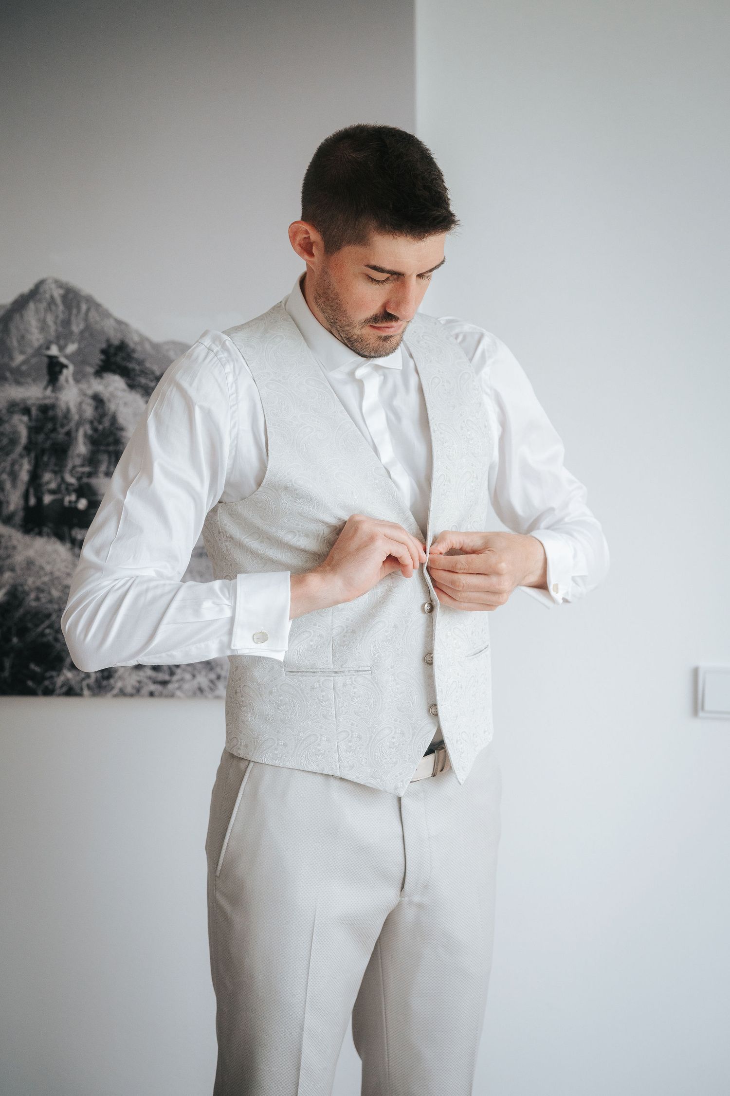 Groom in a white shirt and light gray vest buttons his waistcoat while getting ready indoors beside a framed black-and-white mountain photo.