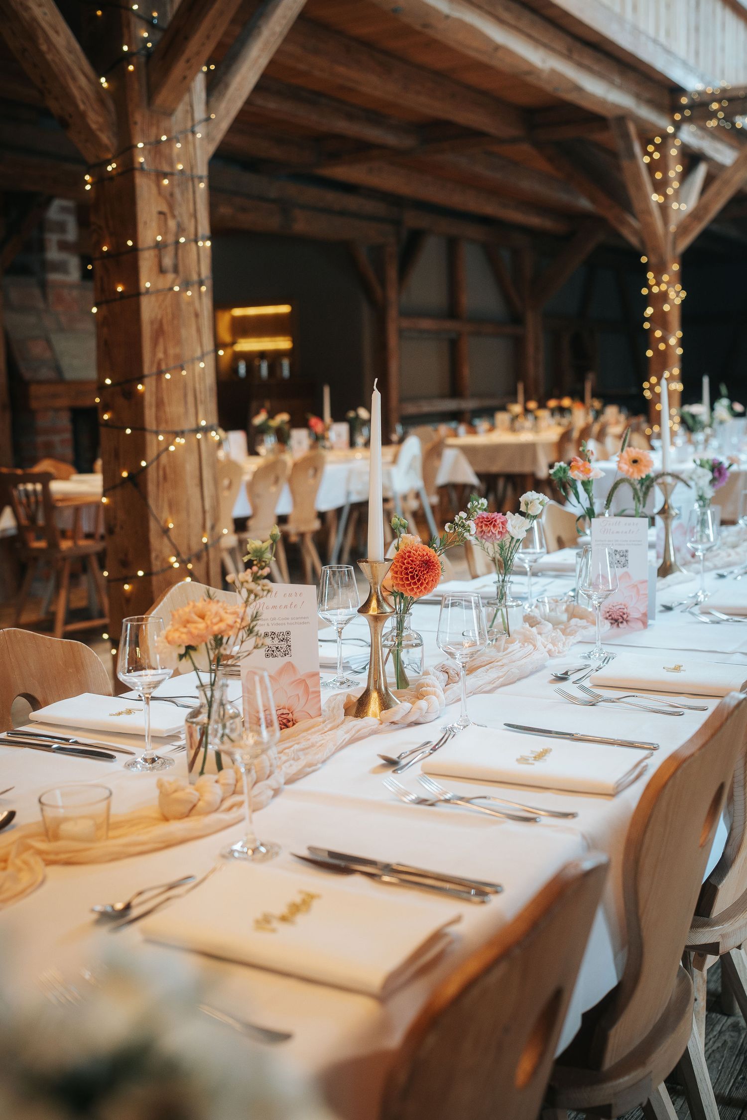 Rustic barn wedding reception with long white-linen tables, wildflower centerpieces, taper candles, and warm string lights on timber beams.