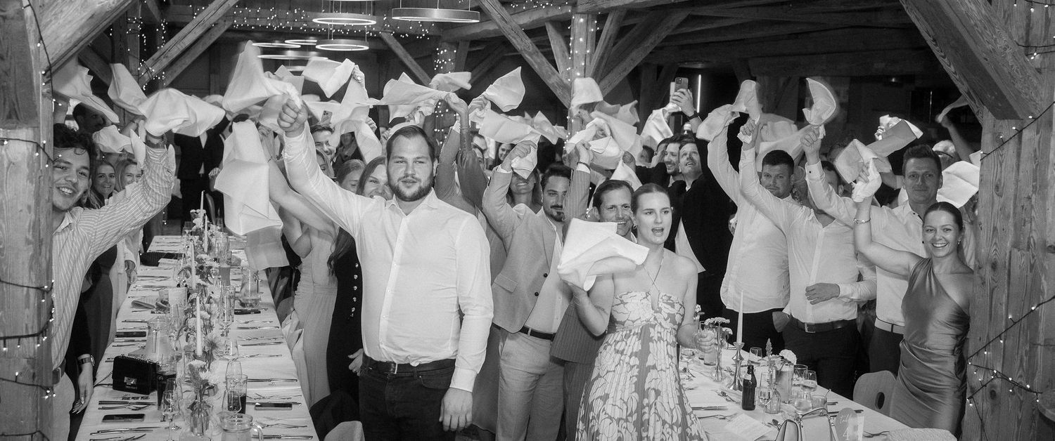 Wedding guests cheer and wave white napkins in the air beside long banquet tables inside a rustic wooden barn venue.
