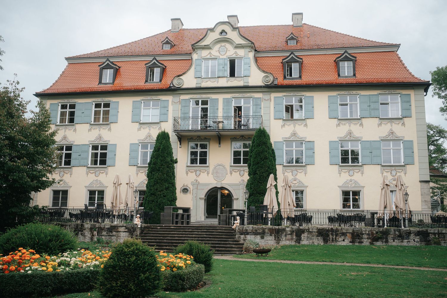 A historic cream manor house with an orange tiled roof and gray shutters, set behind a manicured garden with colorful flowers.