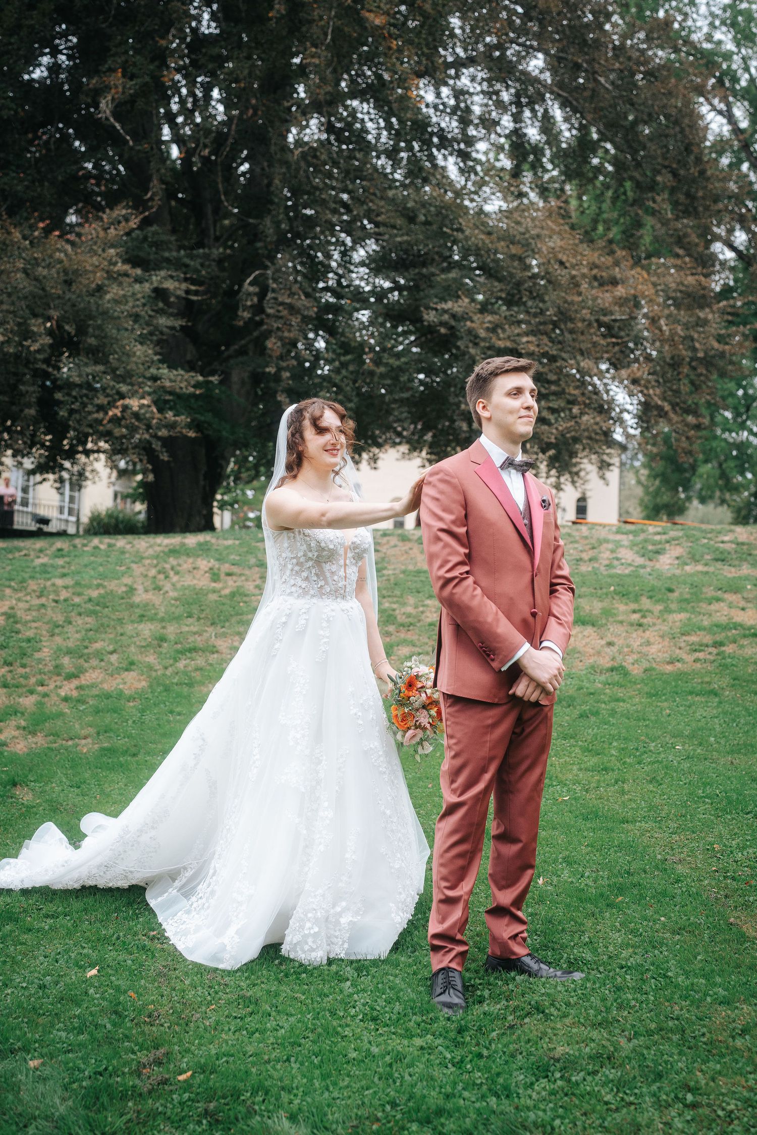 Bride in a white lace gown touches the shoulder of a groom in a rust suit during a first look on a grassy lawn.