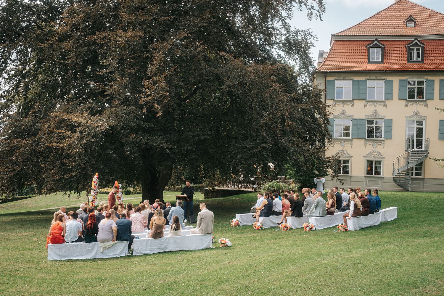 Wedding guests sit on white benches on a sunny lawn beneath a large tree for an outdoor ceremony beside a historic manor house.
