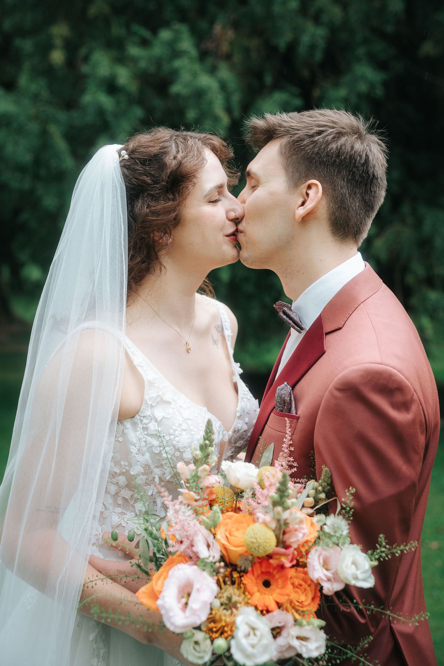 Bride in a lace gown and veil kisses groom in a rust-colored suit, holding an orange and pink bouquet in a green garden.