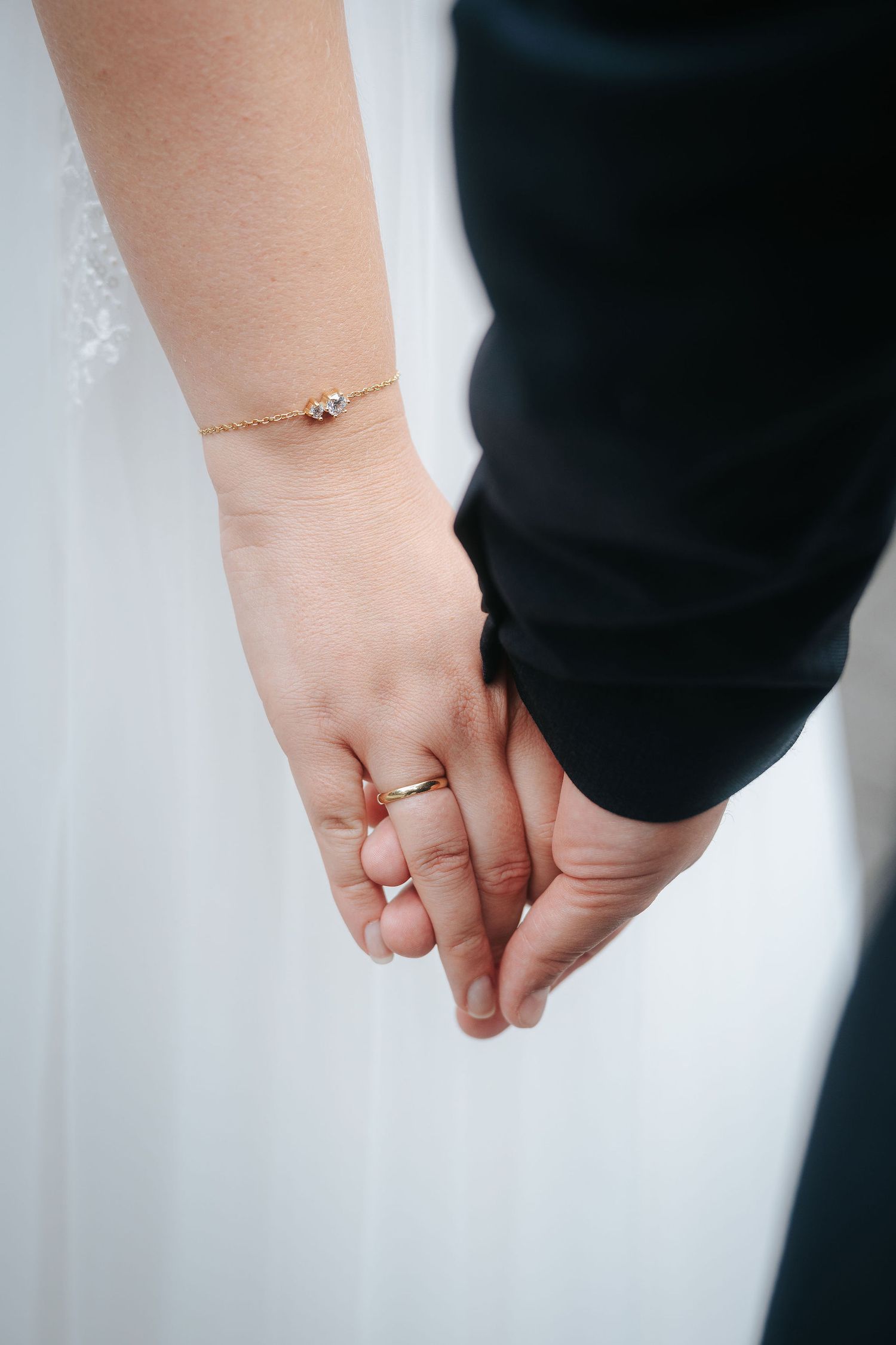 Bride in a white lace dress holding hands with a groom in a dark suit, showing a gold wedding ring and delicate bracelet.