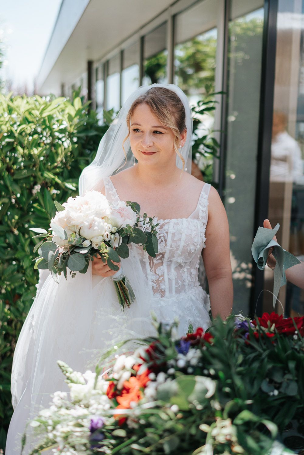 Bride holding bouquet of white flowers on sunny day stands among garden greenery.