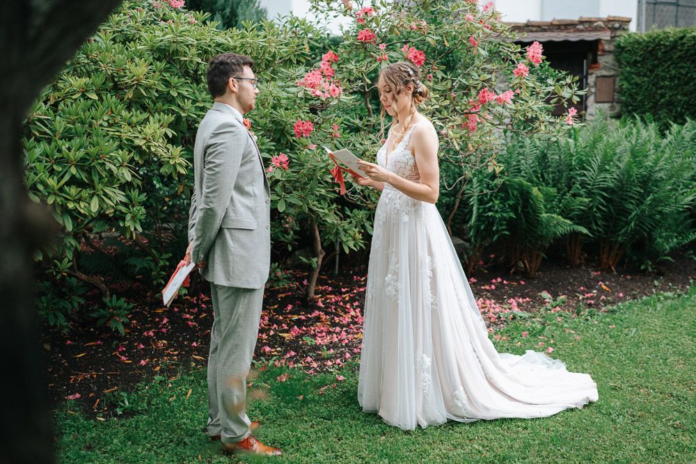 Wedding couple stands in a garden setting with blooming red roses and lush greenery during their first look moment.
