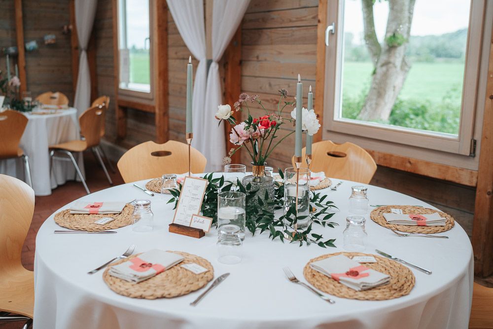 Elegant round dining table set with charger plates, greenery centerpiece, and natural light in rustic wood cabin room.