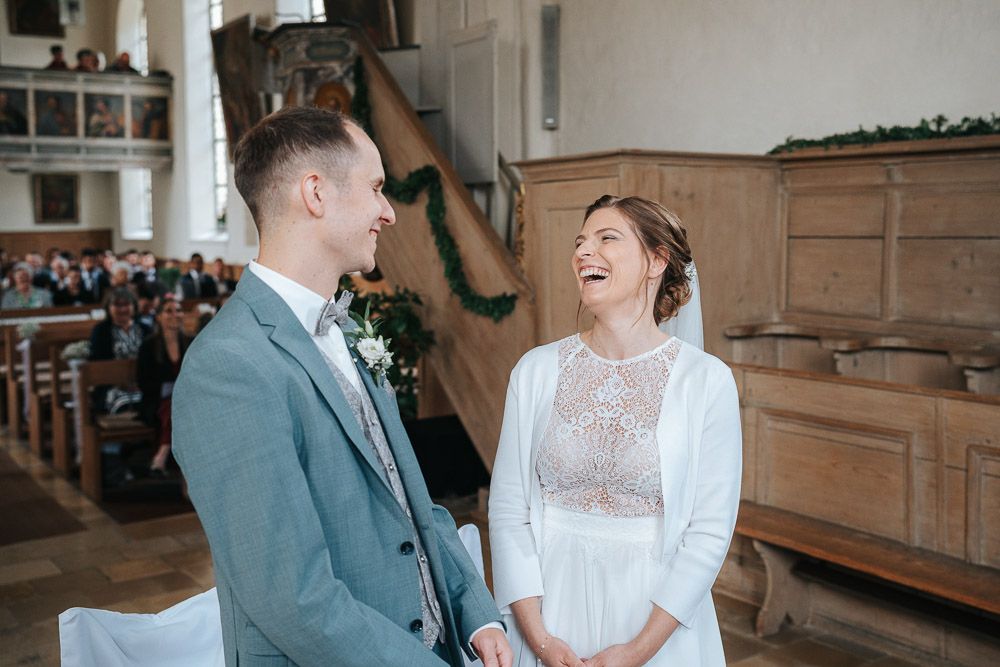 Joyful couple shares a candid laughing moment together during their wedding ceremony in a rustic church setting.