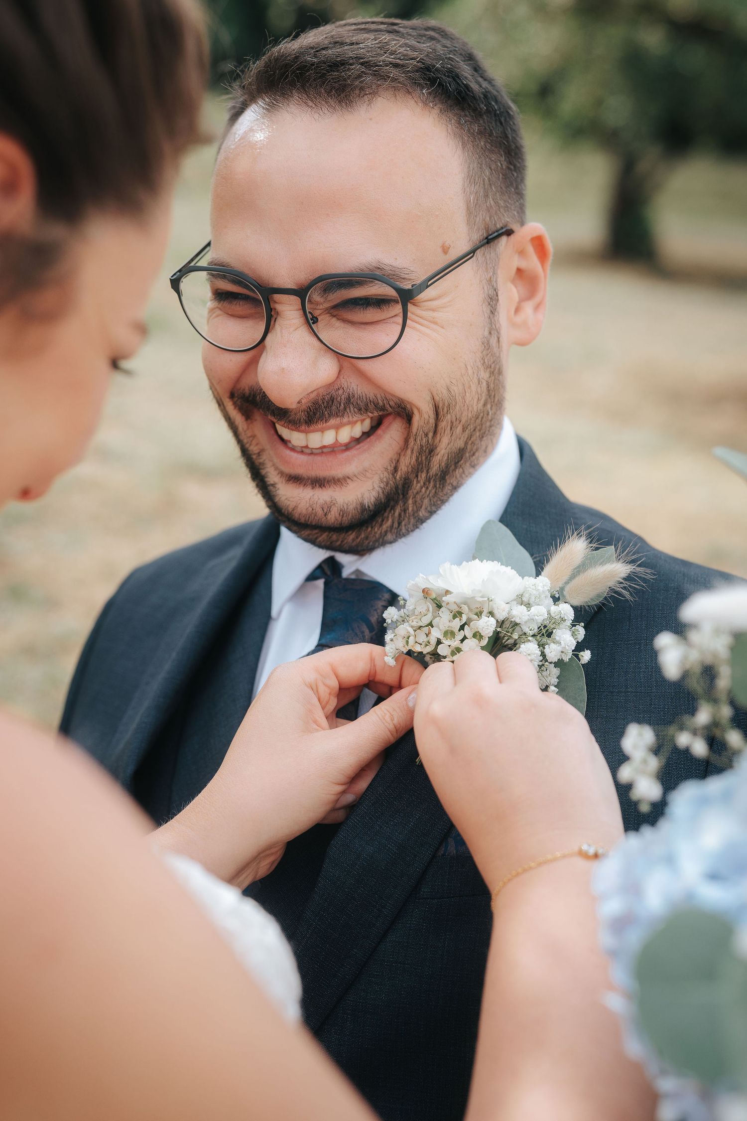 Smiling groom in glasses and a navy suit has a white floral boutonniere pinned to his lapel by a bride at an outdoor wedding.