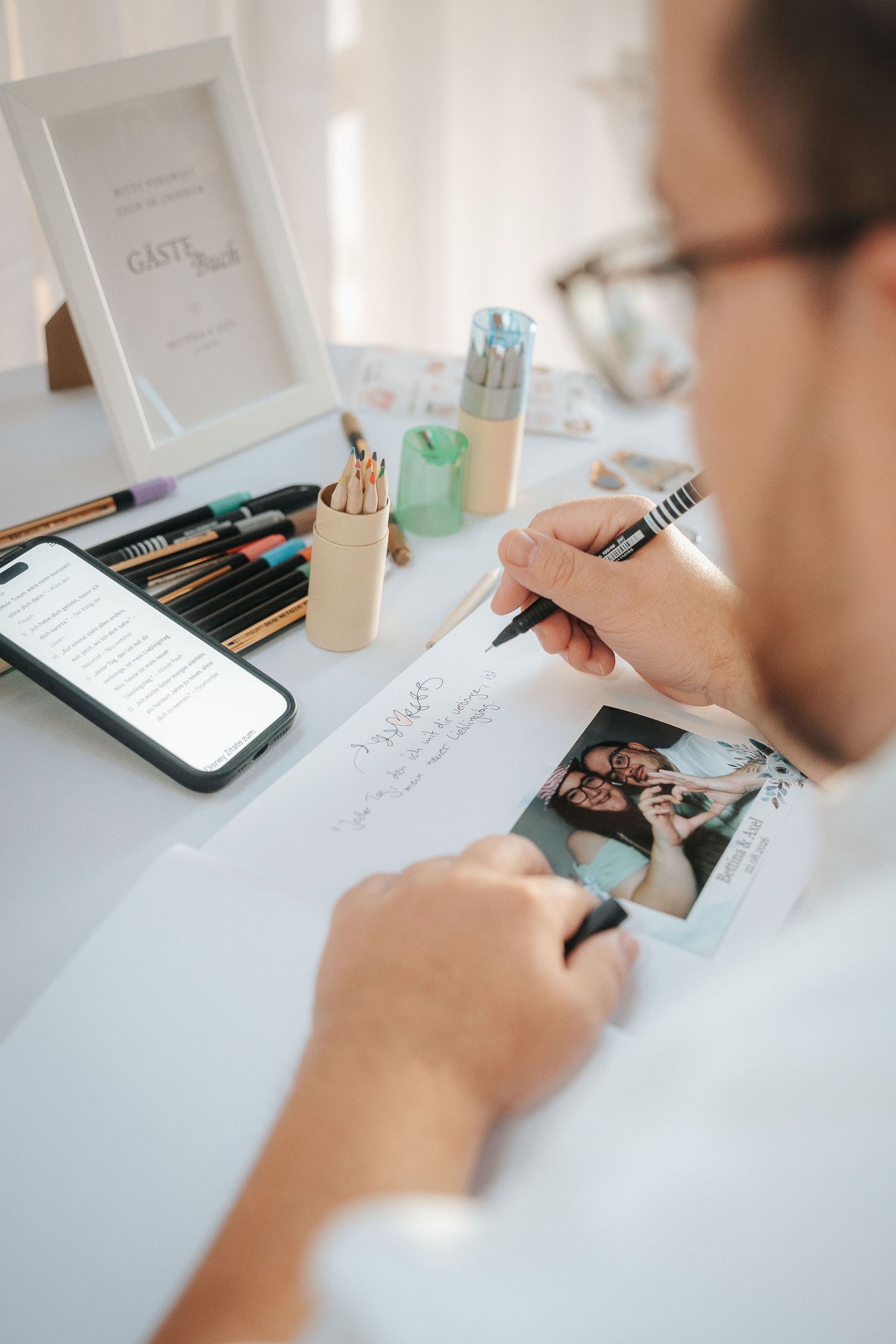 Man with glasses writes a message in a wedding guest book beside an instant photo, colored pencils, pens and a smartphone on a table.