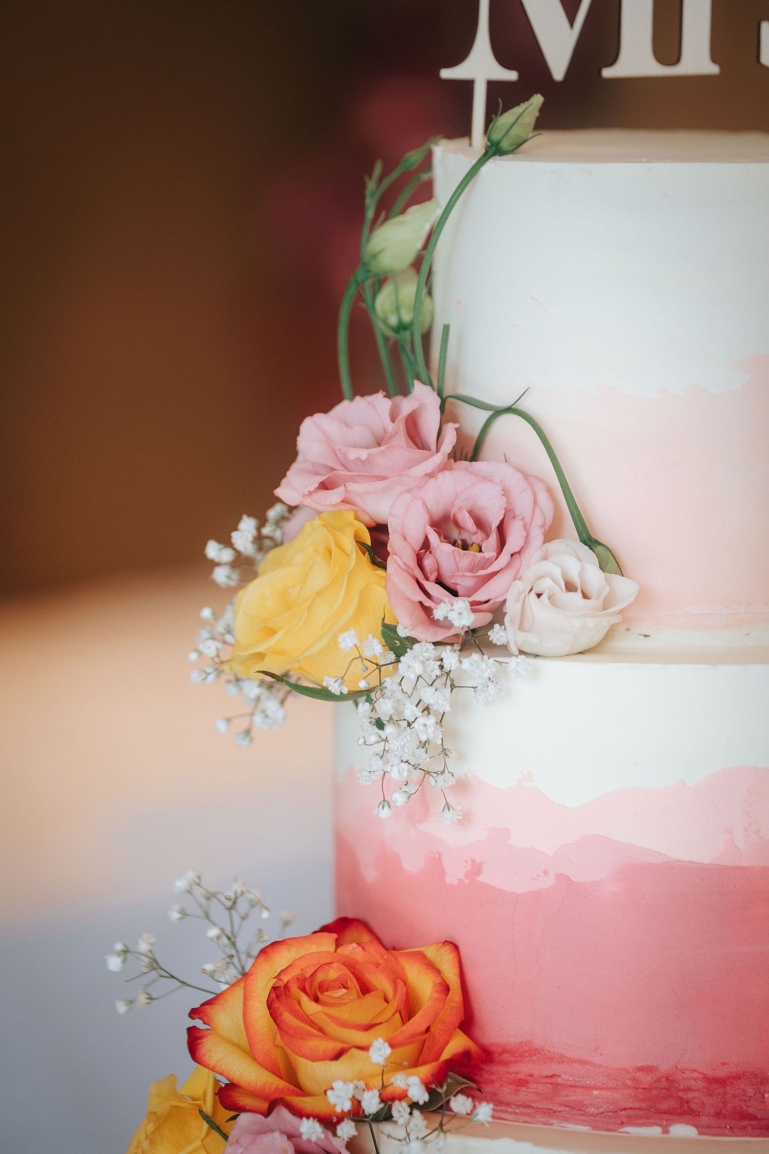 Tiered wedding cake with pink watercolor ombre frosting, decorated with fresh roses, baby's breath, and a cake topper in warm soft light.