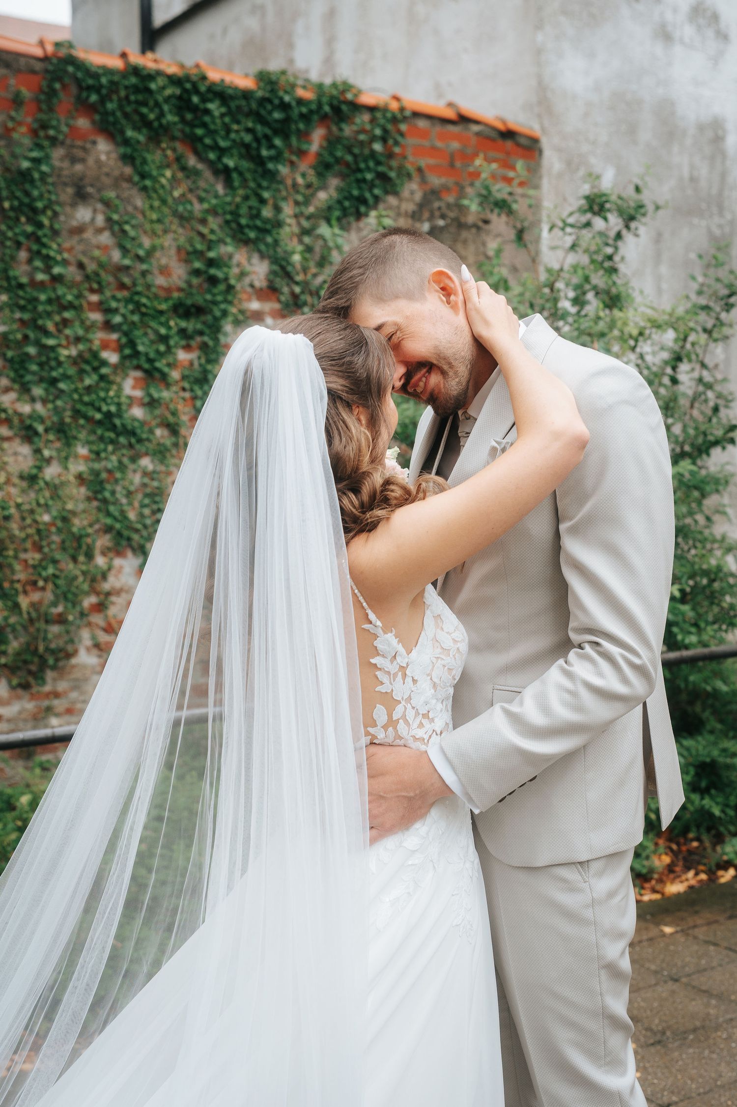Bride in a lace gown and long veil embraces a smiling groom in a beige suit beside an ivy-covered brick wall.