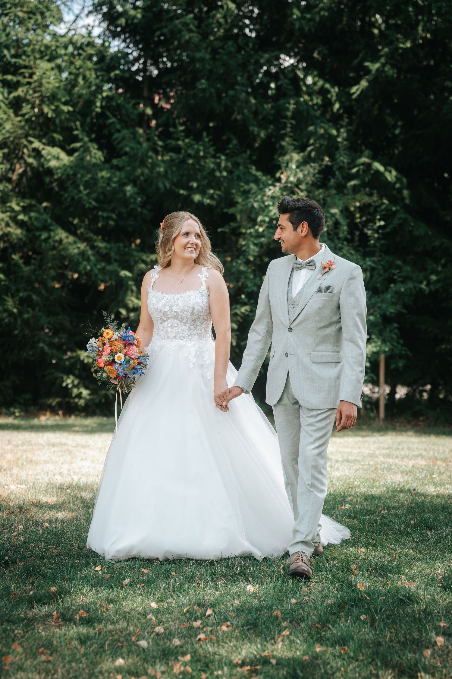 Bride in a white lace gown holding a colorful bouquet walks hand in hand with a groom in a light gray suit across a sunny green lawn.