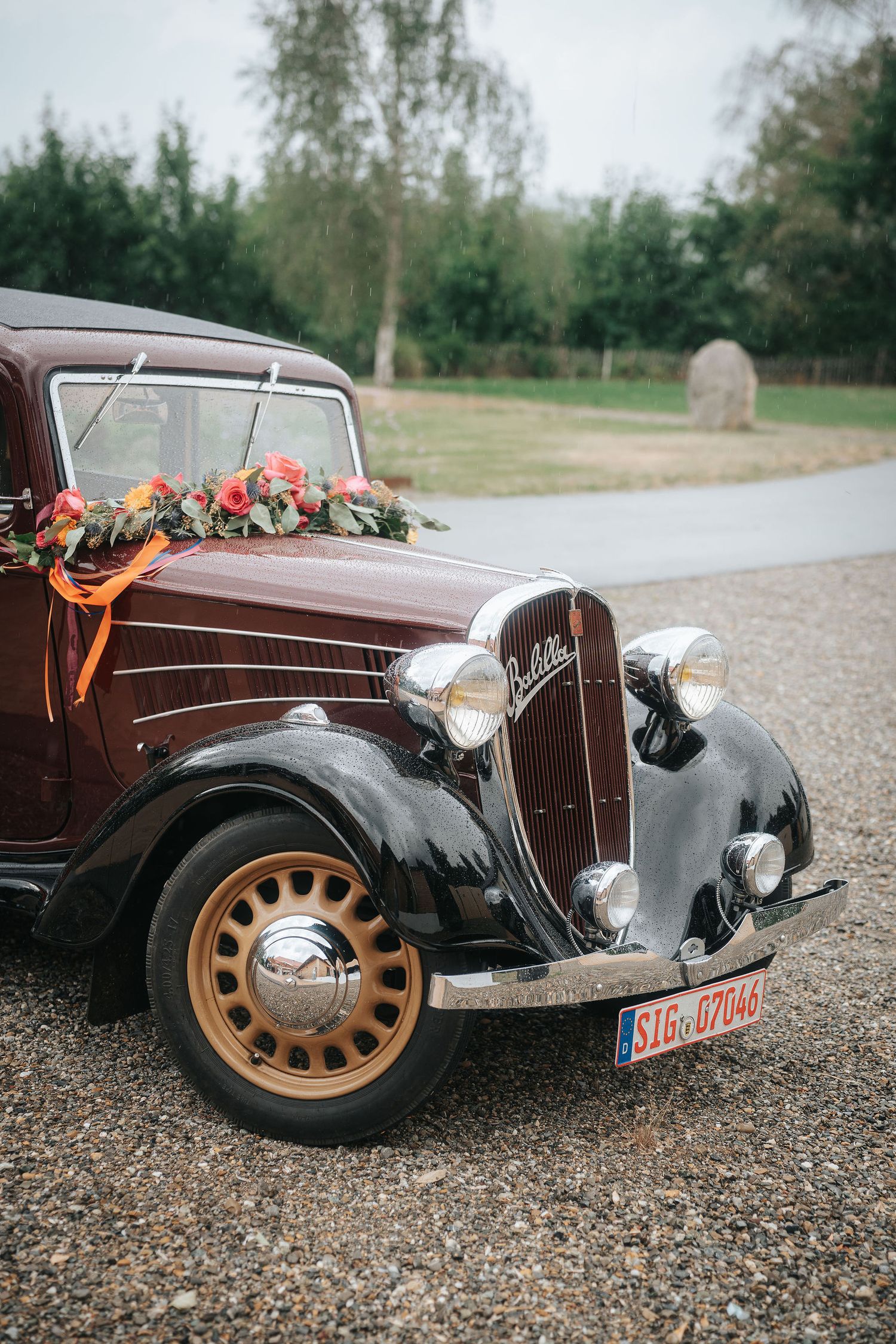 Vintage dark red wedding car with orange wheels and a floral garland of pink and orange flowers on the hood, parked on a gravel drive.