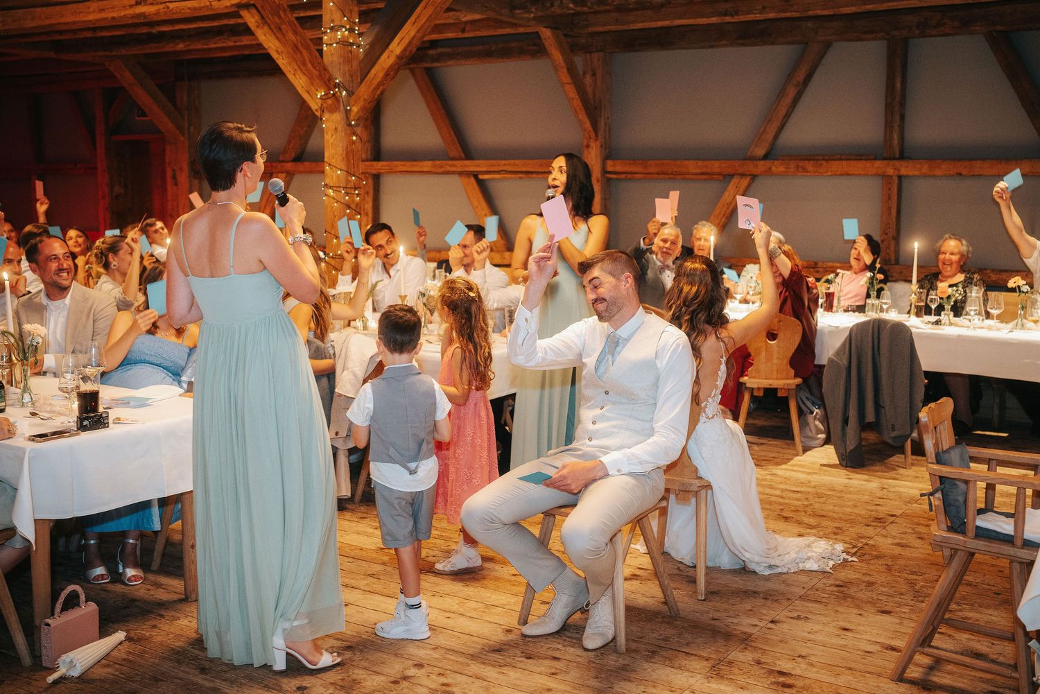 Groom in a white shirt seated on a chair holding up a paper sign while wedding guests laugh in a rustic wooden barn reception.