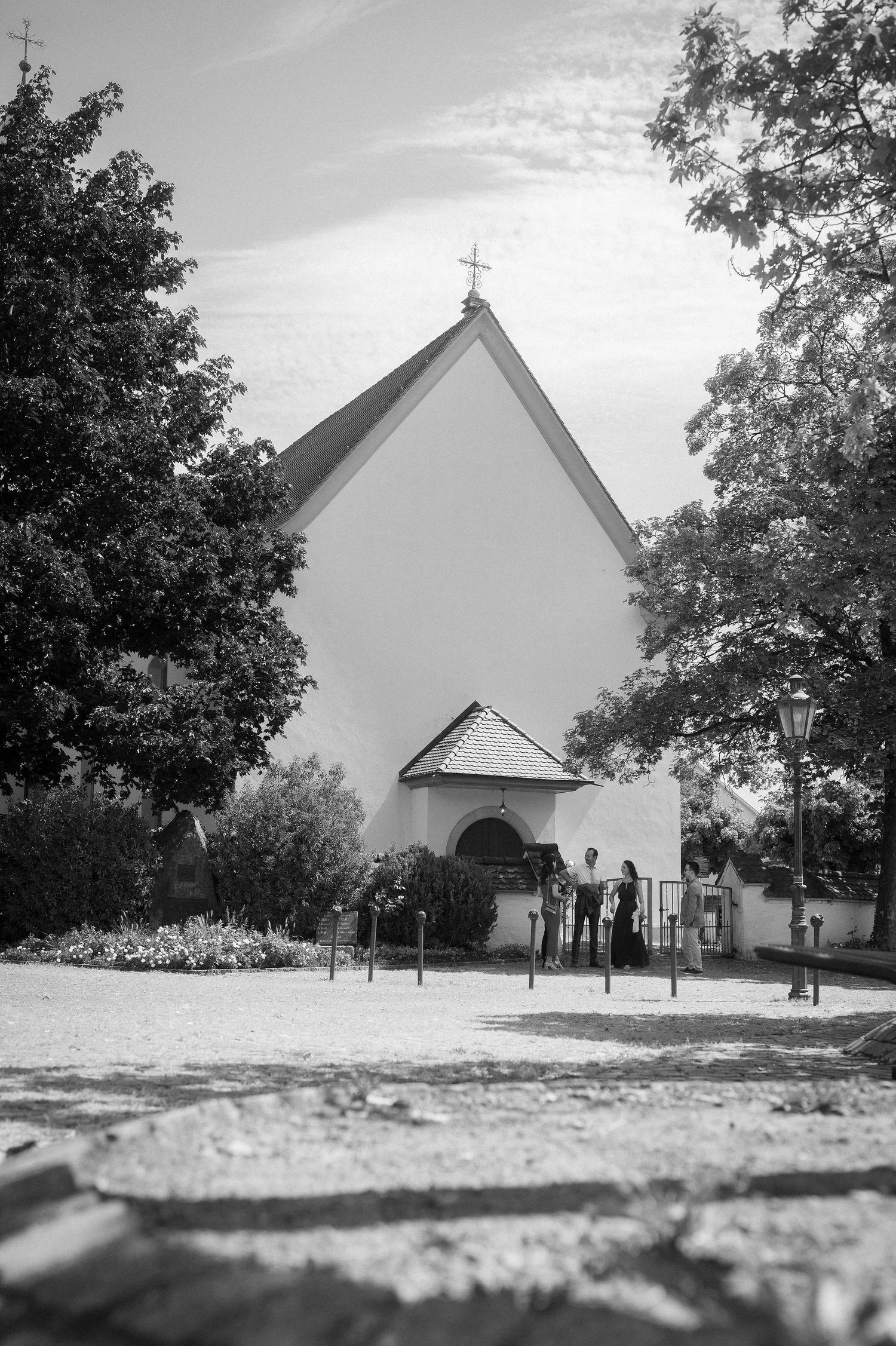 Black and white photo of a white church with a cross on the roof, surrounded by trees on a sunny day.