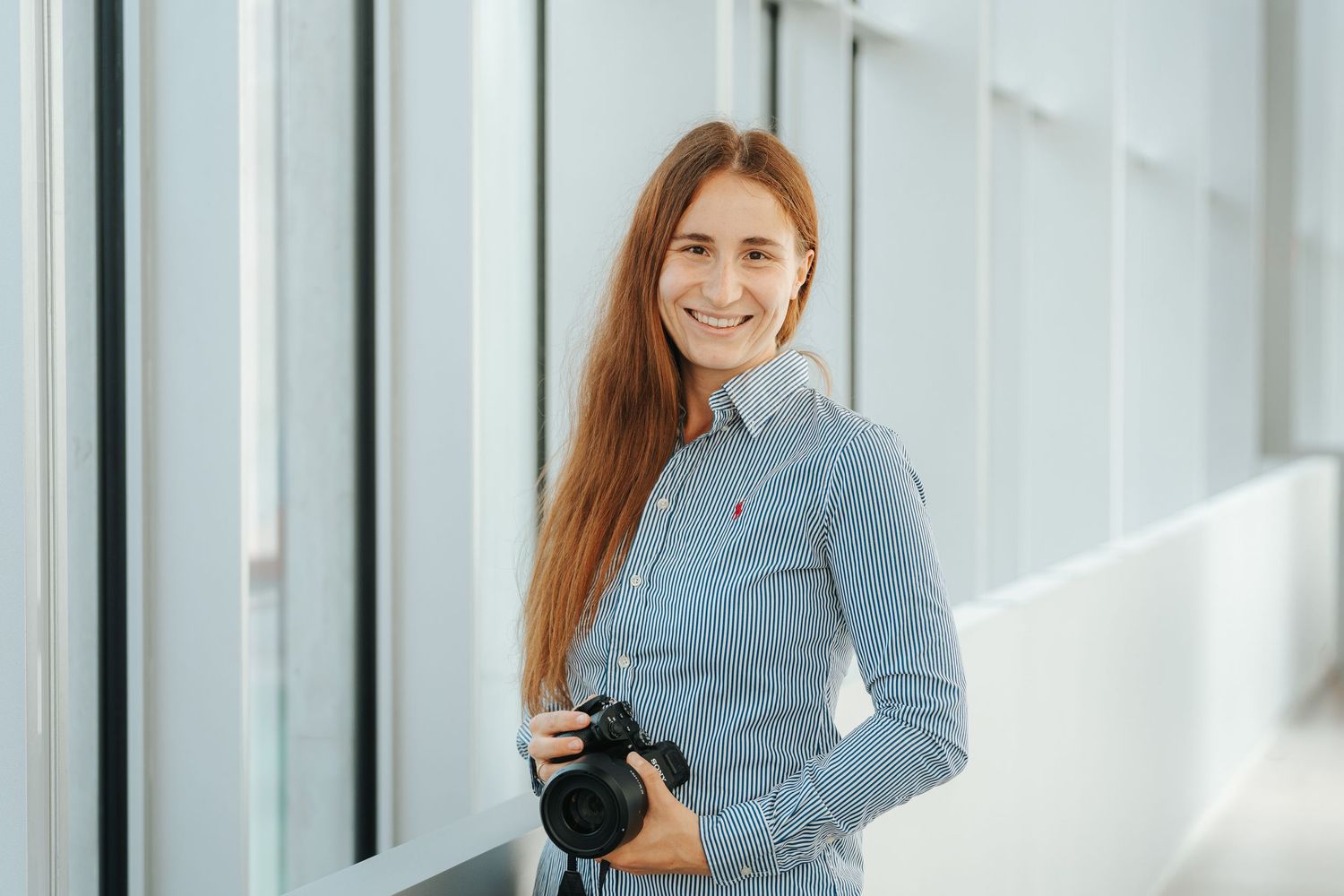 Professional woman in blue blazer stands confidently in bright modern office hallway with glass walls.