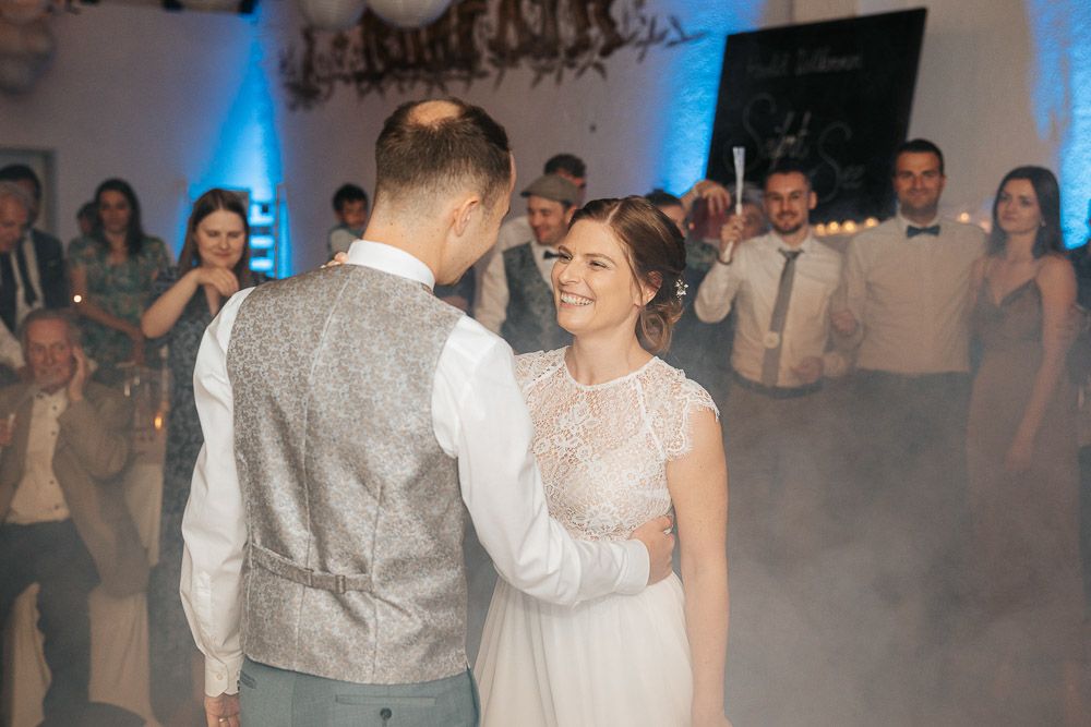 A couple shares their first dance as newlyweds on a misty dance floor illuminated by blue lighting at a wedding reception.