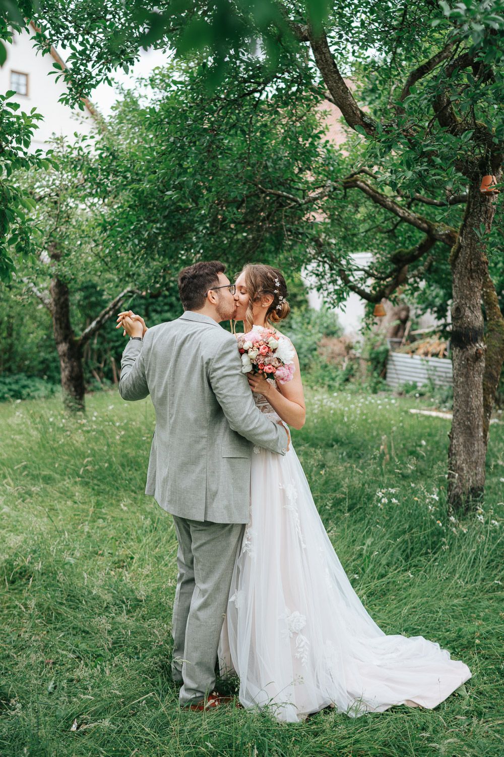 Newlyweds embrace in romantic outdoor garden wedding setting surrounded by lush green trees.
