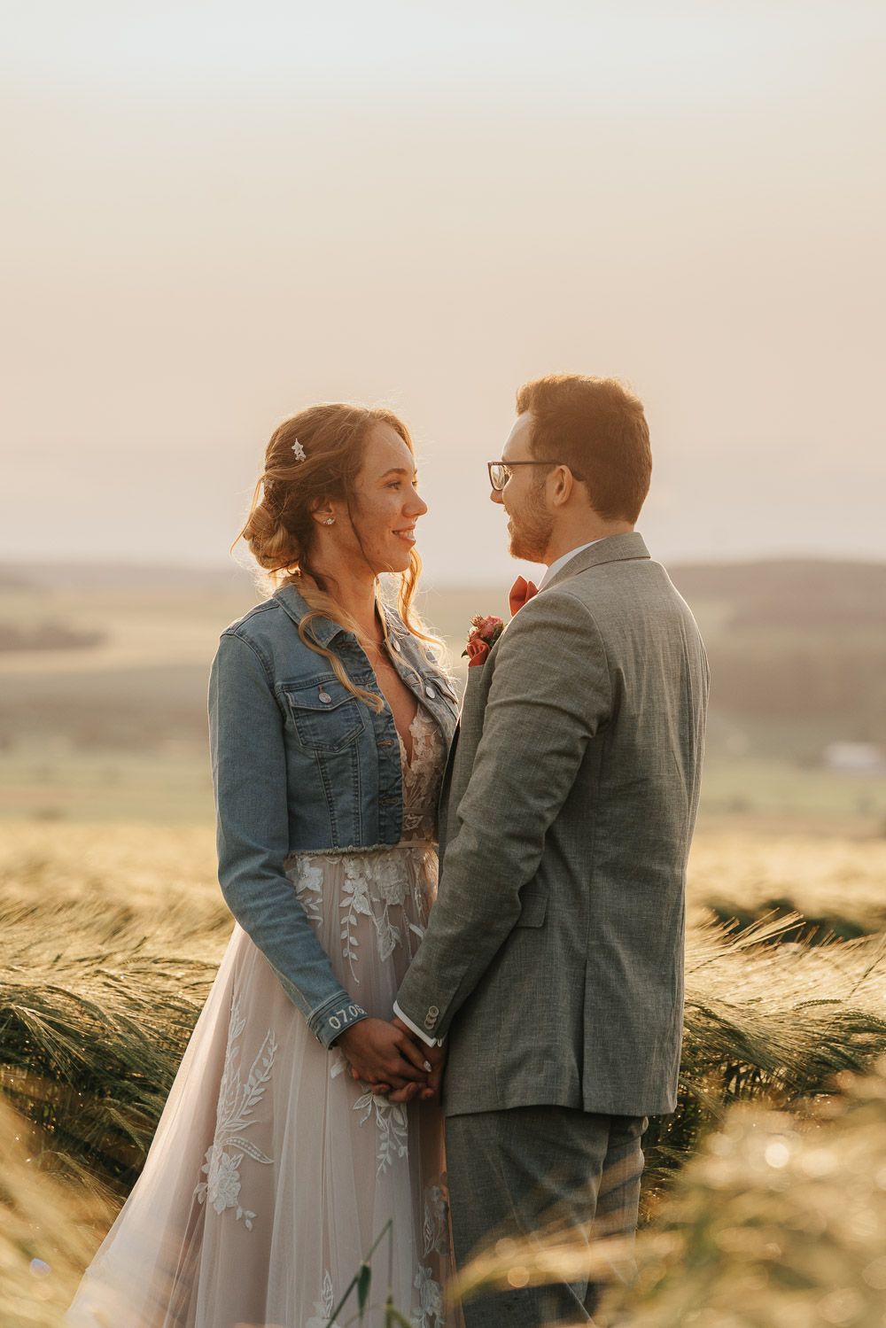 Couple embraces in golden wheat field at sunset wearing denim jacket and suit during romantic outdoor photoshoot.