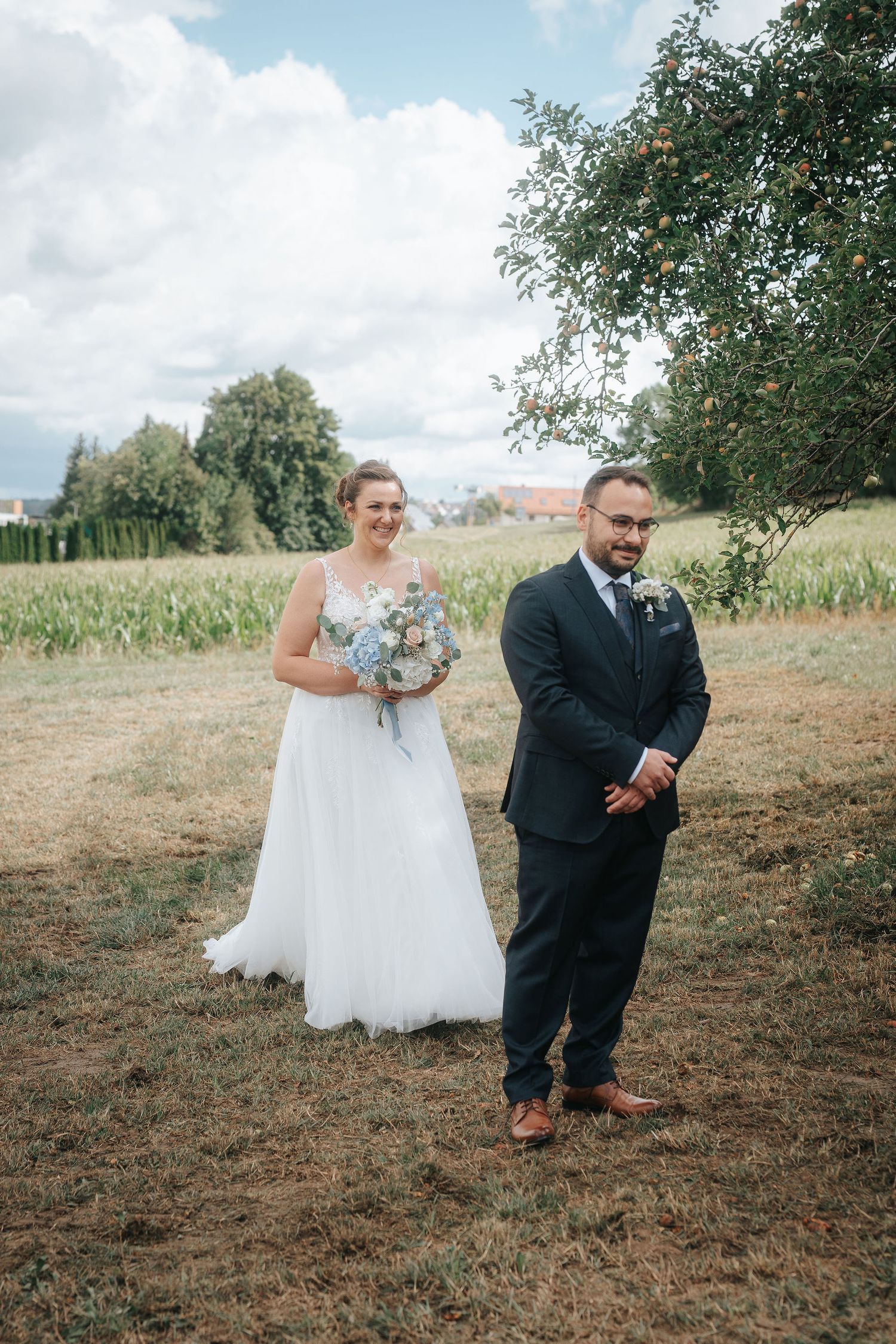 Bride in a white gown holding a blue bouquet smiles behind a groom in a navy suit in a sunny field with an apple tree.