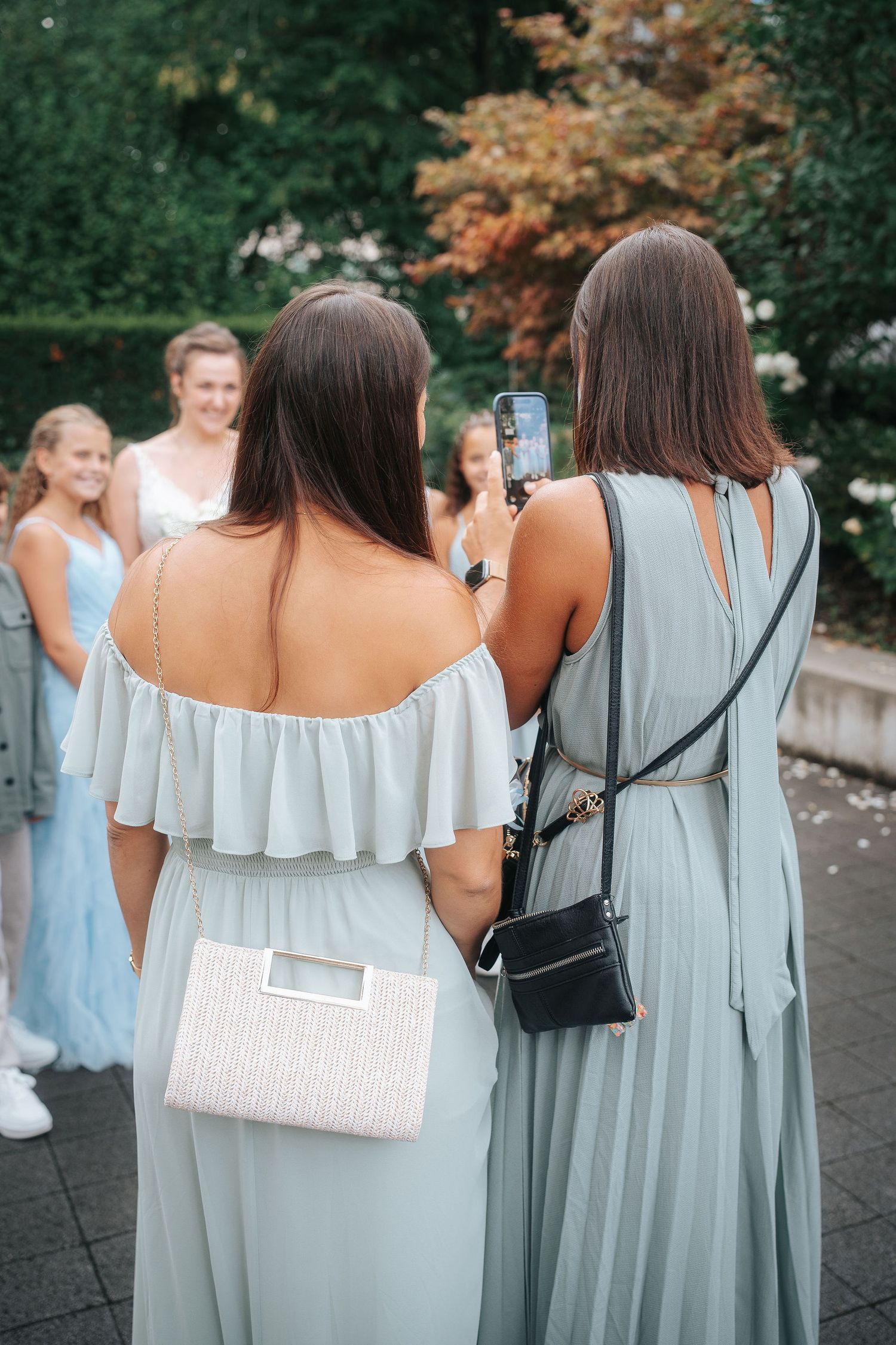 Two bridesmaids in sage green dresses, one holding a woven clutch, stand outdoors while smiling guests gather near a garden.