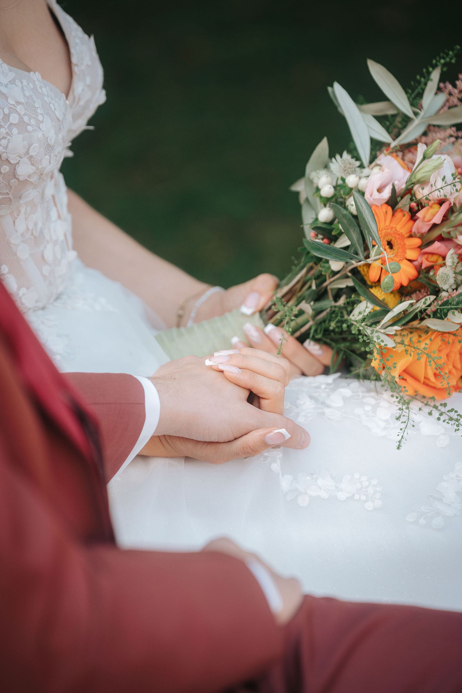 Bride in a white lace wedding dress holds hands with groom in a burgundy suit beside a bouquet of orange flowers and greenery.