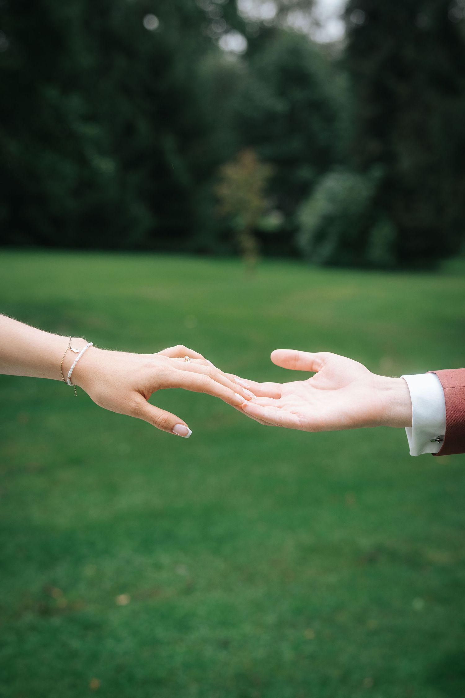 Two hands reaching toward each other over a green lawn, one wearing delicate bracelets and the other in a white shirt cuff.