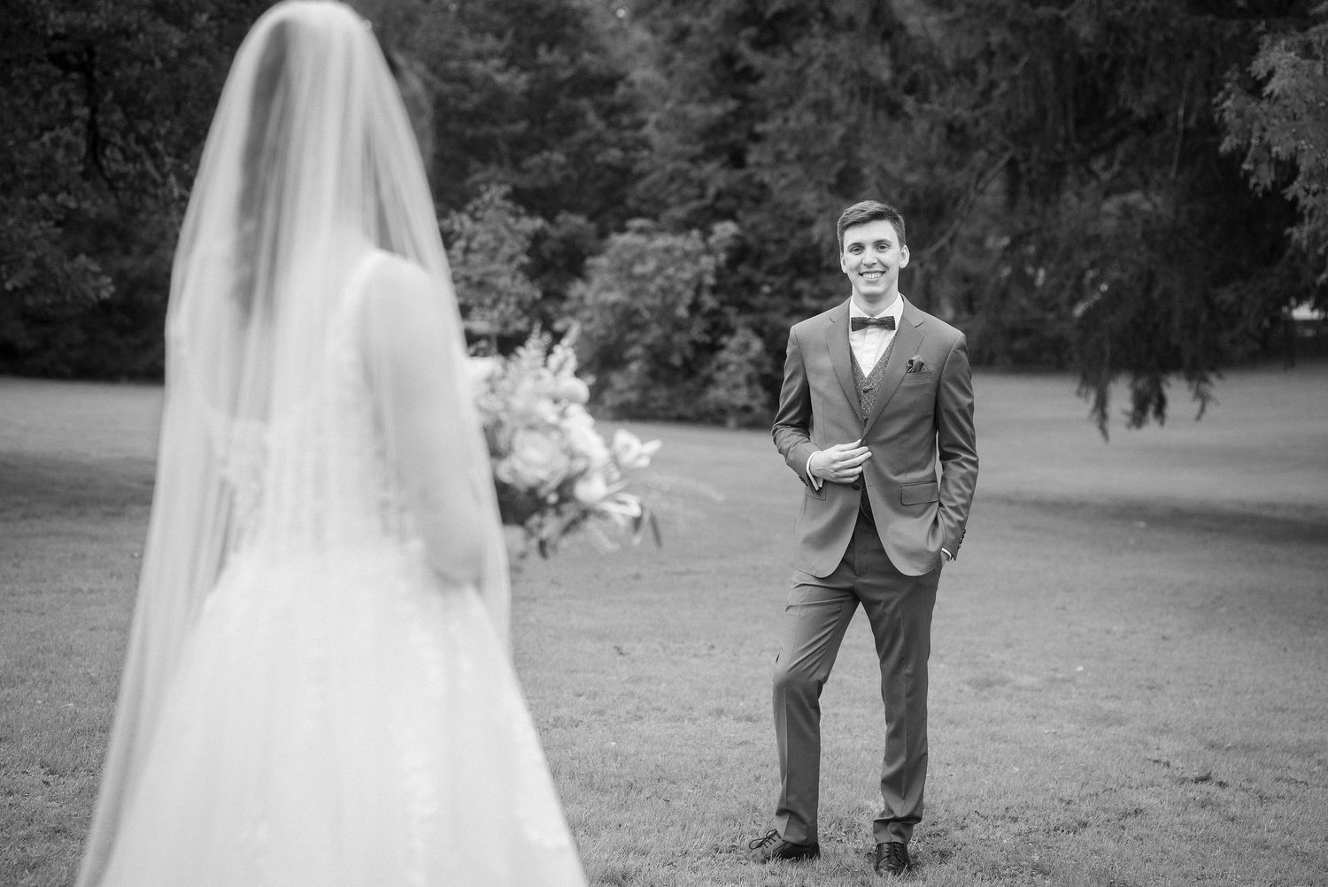 Black and white wedding photo of a smiling groom in a suit and bow tie watching his veiled bride holding a bouquet in a park.